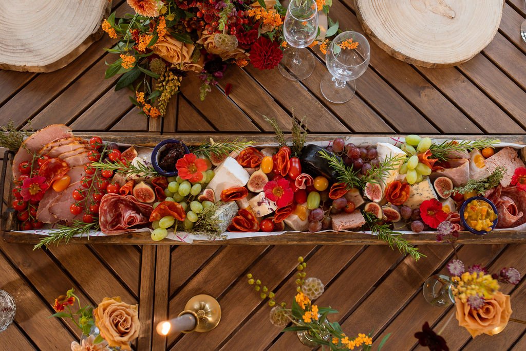 Charcuterie board with cured meats, cheeses, grapes, cherry tomatoes, figs, and herbs on a wooden table with floral decorations.