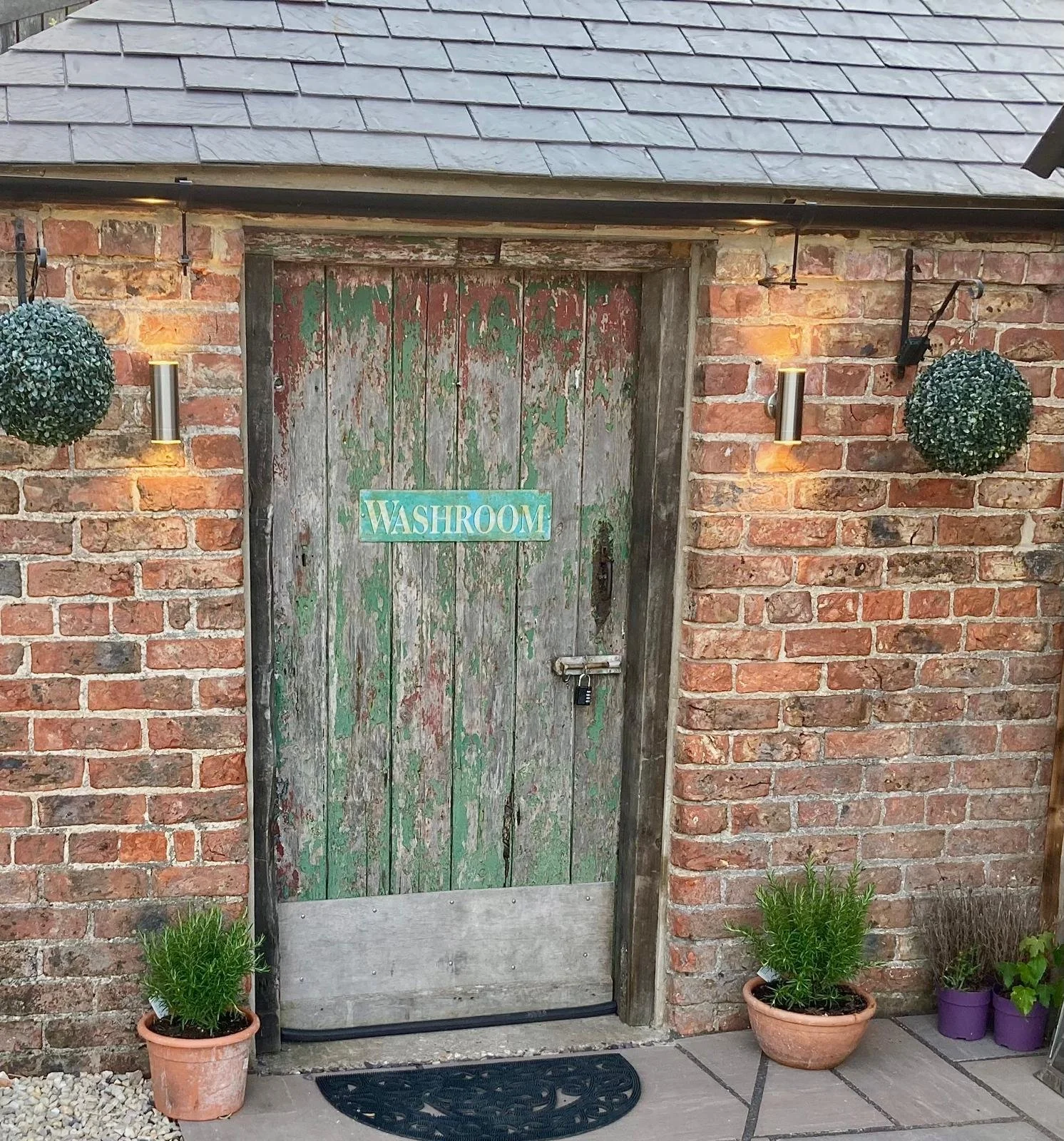 The image shows a rustic wooden door with a chipped green paint finish mounted in a brick wall. Above the door, there's a sloped roof with shingles. On each side of the door, there are outdoor wall-mounted lights and hanging decorative topiary balls.
