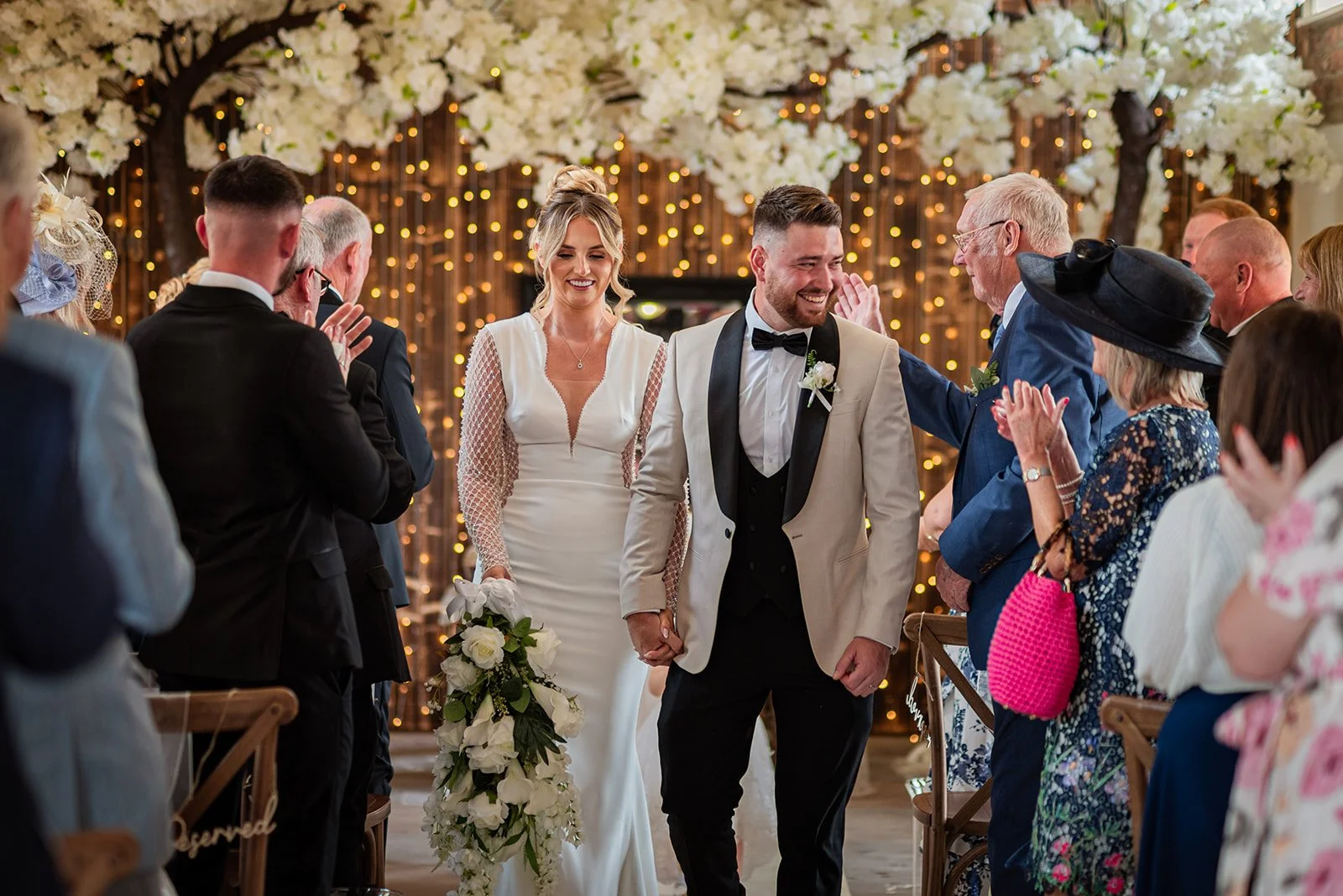 A newlywed couple walking down the aisle after their wedding ceremony, surrounded by family and friends clapping and celebrating indoors with a decorated background of white blossoms and warm string lights.