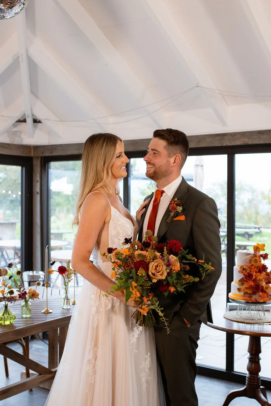 A bride and groom smiling at each other indoors during their wedding. The bride is holding a large bouquet of colorful flowers and wearing a lace wedding gown. The groom is dressed in a dark suit with a white shirt, orange tie, and boutonniere. There are flowers and a tiered wedding cake decorated with orange and red flowers in the background.