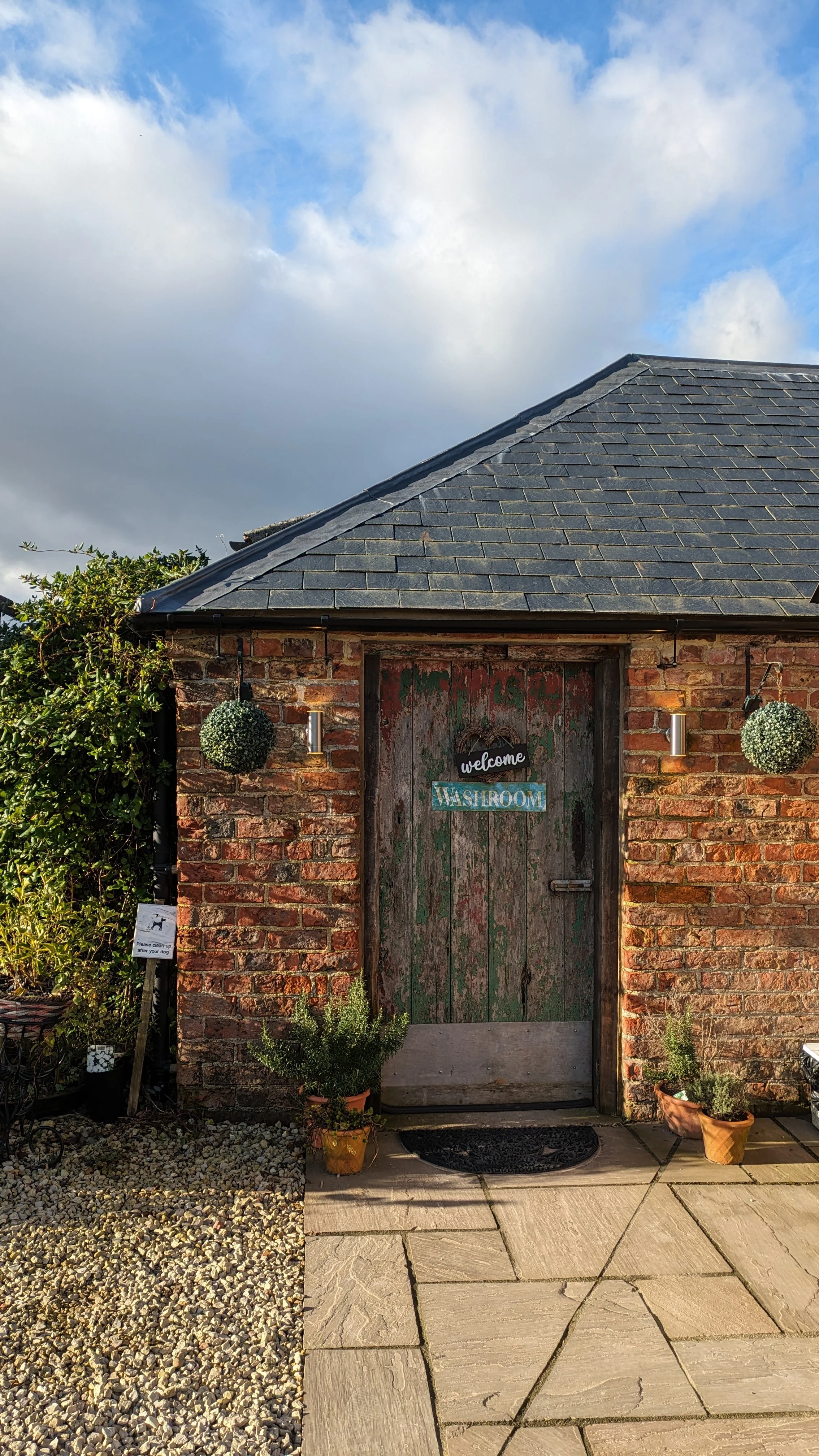 Rustic brick building with a weathered green wooden door labeled 'welcome' and 'WASHROOM.' There are two round hanging plants, two wall-mounted lights, potted plants on either side of the door, and outdoor paving with a small gravel section to the le