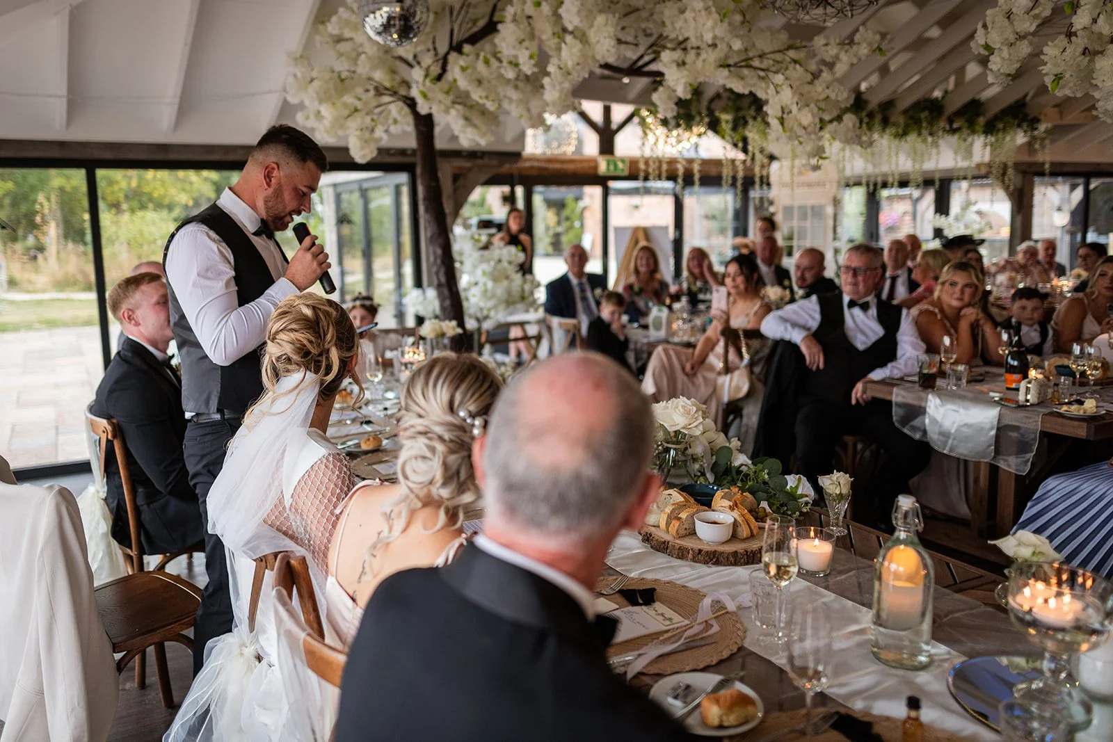 A wedding reception with a man giving a speech using a microphone, seated at a table with a bride and groom, surrounded by guests sitting at decorated tables with flowers, candles, and food.
