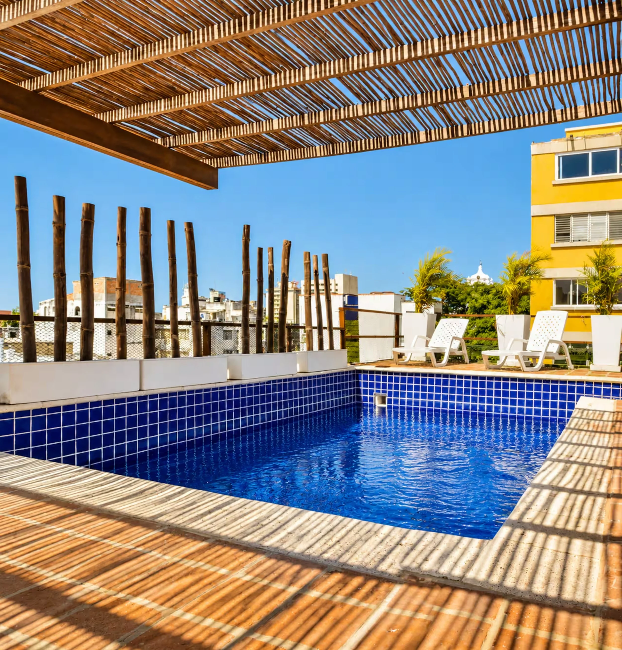 View of outdoor swimming pool with blue tiles, surrounded by a wooden pergola structure, poolside white chairs, and a yellow building under a clear blue sky.