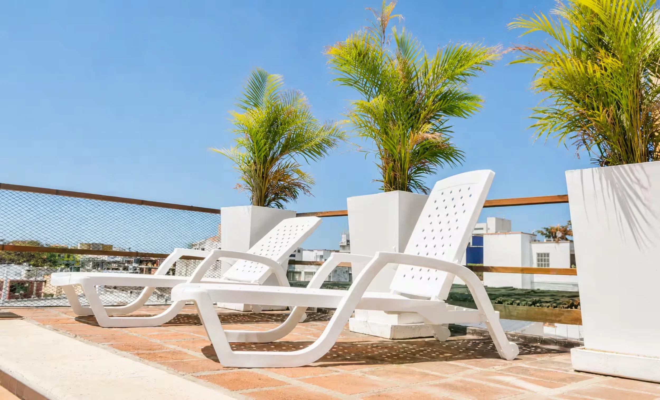 Rooftop with white lounge chairs, green potted plants, roof railing, and building skyline under clear blue sky.