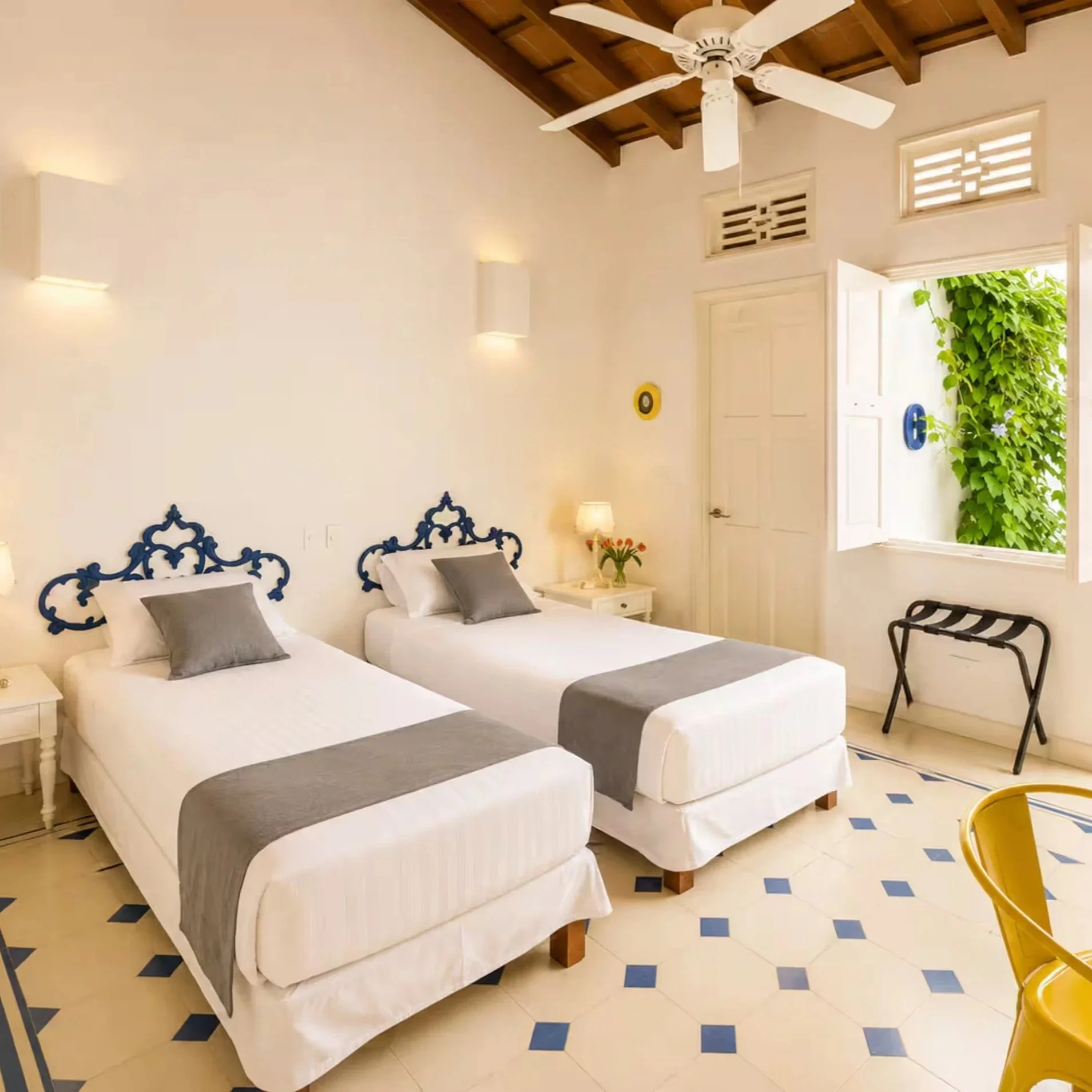 Room with three beds, white bed linens, dark blue headboards, gray pillows, small white nightstand with a lamp and flowers, open window with greenery outside, white walls, wood ceiling with a ceiling fan, tiled floor with a blue diamond pattern, and a yellow chair.