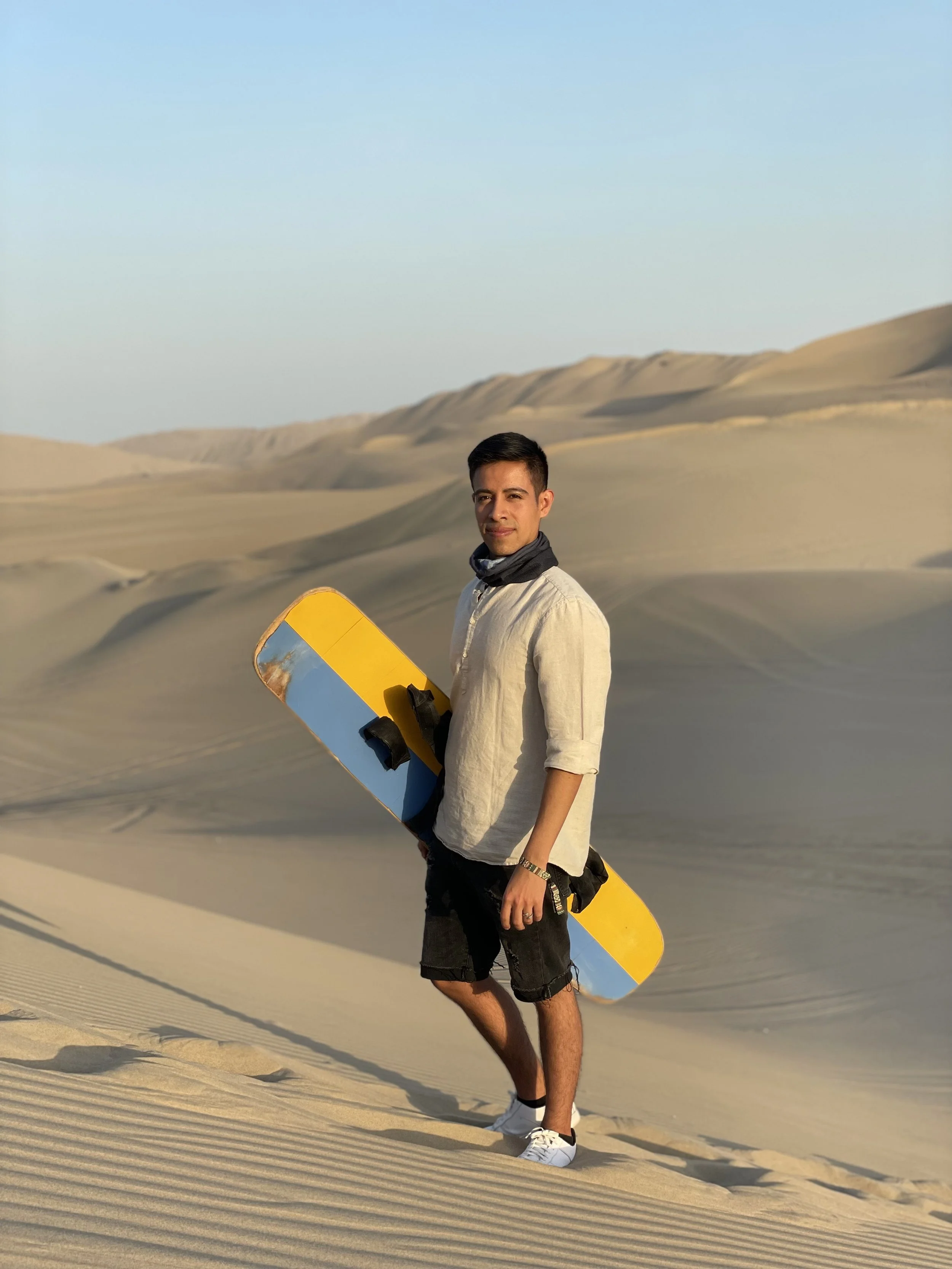 A man standing in a desert with sand dunes, holding a skateboard, wearing a beige shirt, black shorts, and white sneakers.