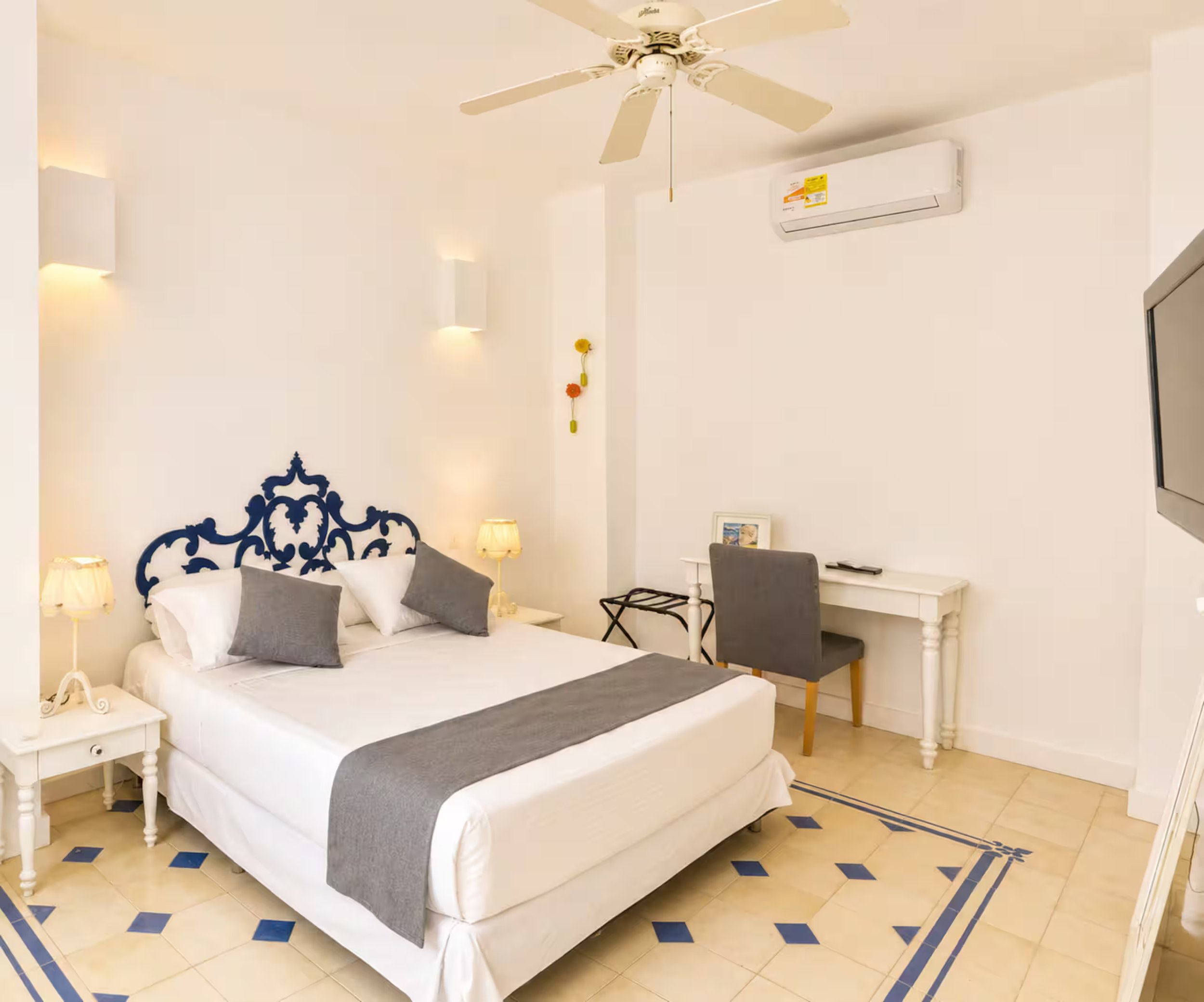 A bedroom with a white bed featuring gray and white pillows, a white bedside table with a lamp, a wall-mounted air conditioning unit, a decorative wall hanging with flowers, and a corner with a small table and chair. The room has beige tiled floor wi