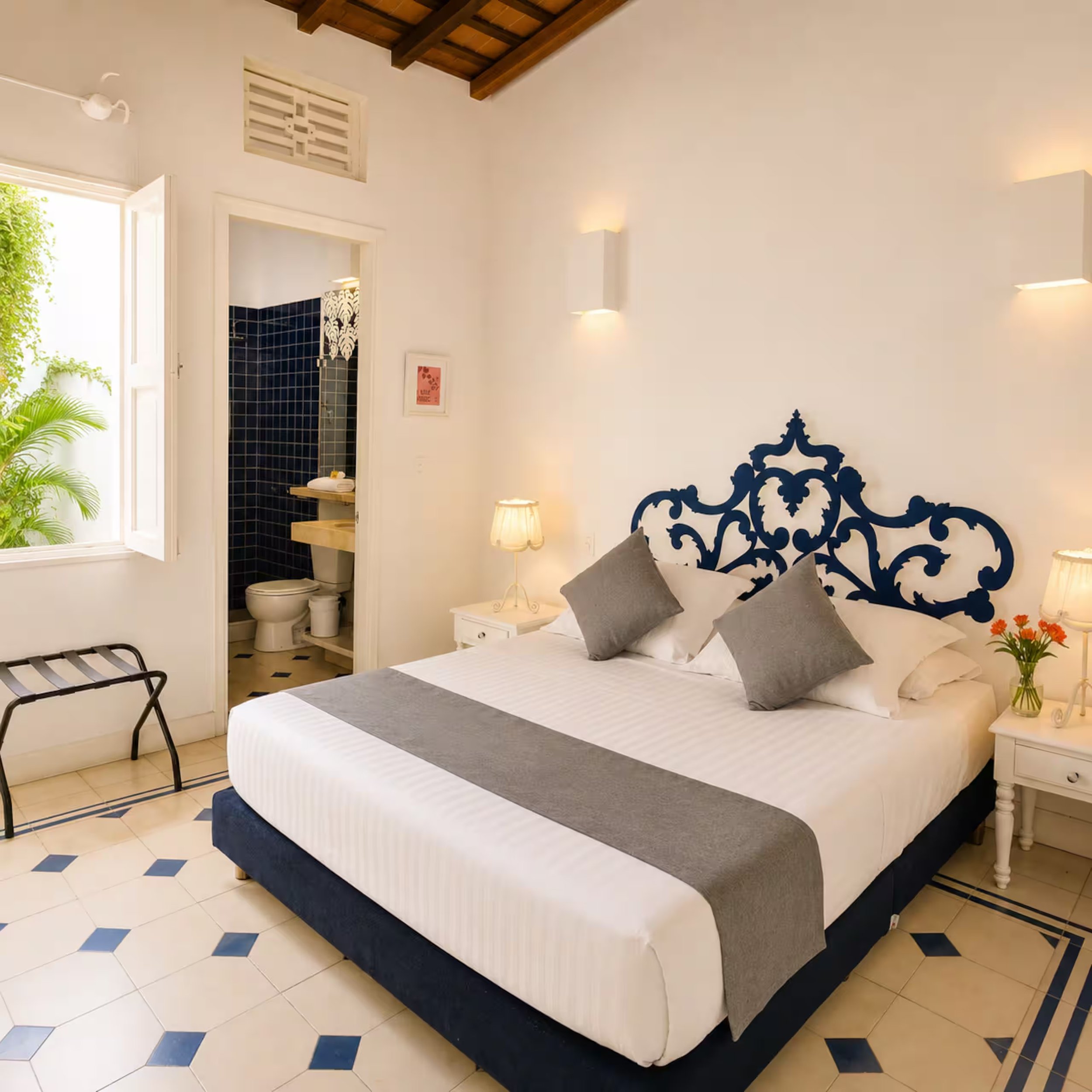 A bedroom with a bed that has a black and white headboard, gray pillows, and white bedding with a gray runner. There is a small white nightstand with a lamp and flowers on it. The wall behind the bed has two modern wall sconces and a small framed pic