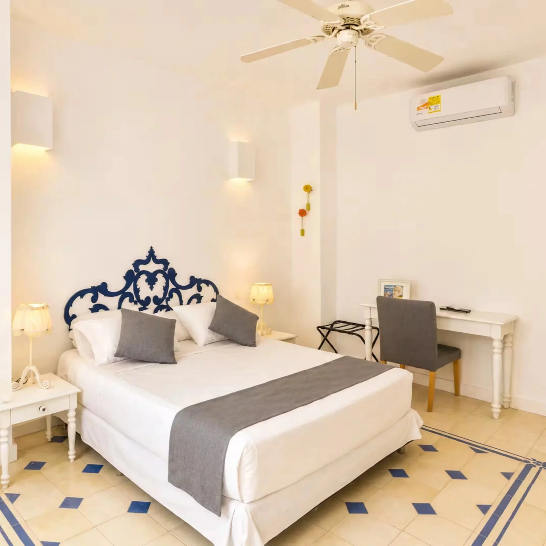 A bedroom with white walls and a ceiling fan. The bed has a white frame, white bedding, and gray pillows. There are two small white nightstands with lamps, a gray chair, a small white table, and a white dresser with a framed picture. The floor is tiled with beige and blue patterns.
