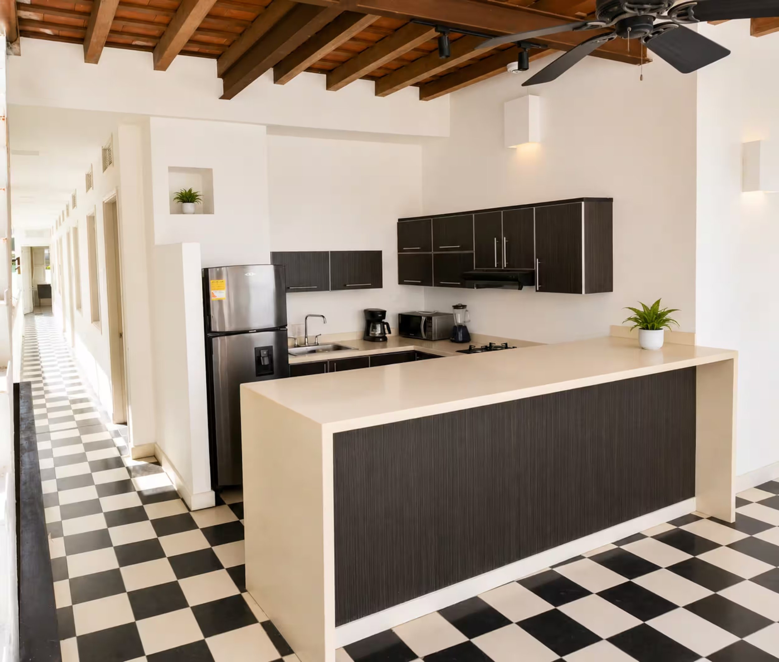 Kitchen with dark cabinets, a stainless steel refrigerator, a coffee maker, and a microwave on a beige countertop. There are plants on a shelf and on the island. The ceiling has exposed wooden beams and a ceiling fan. The floor is black and white che