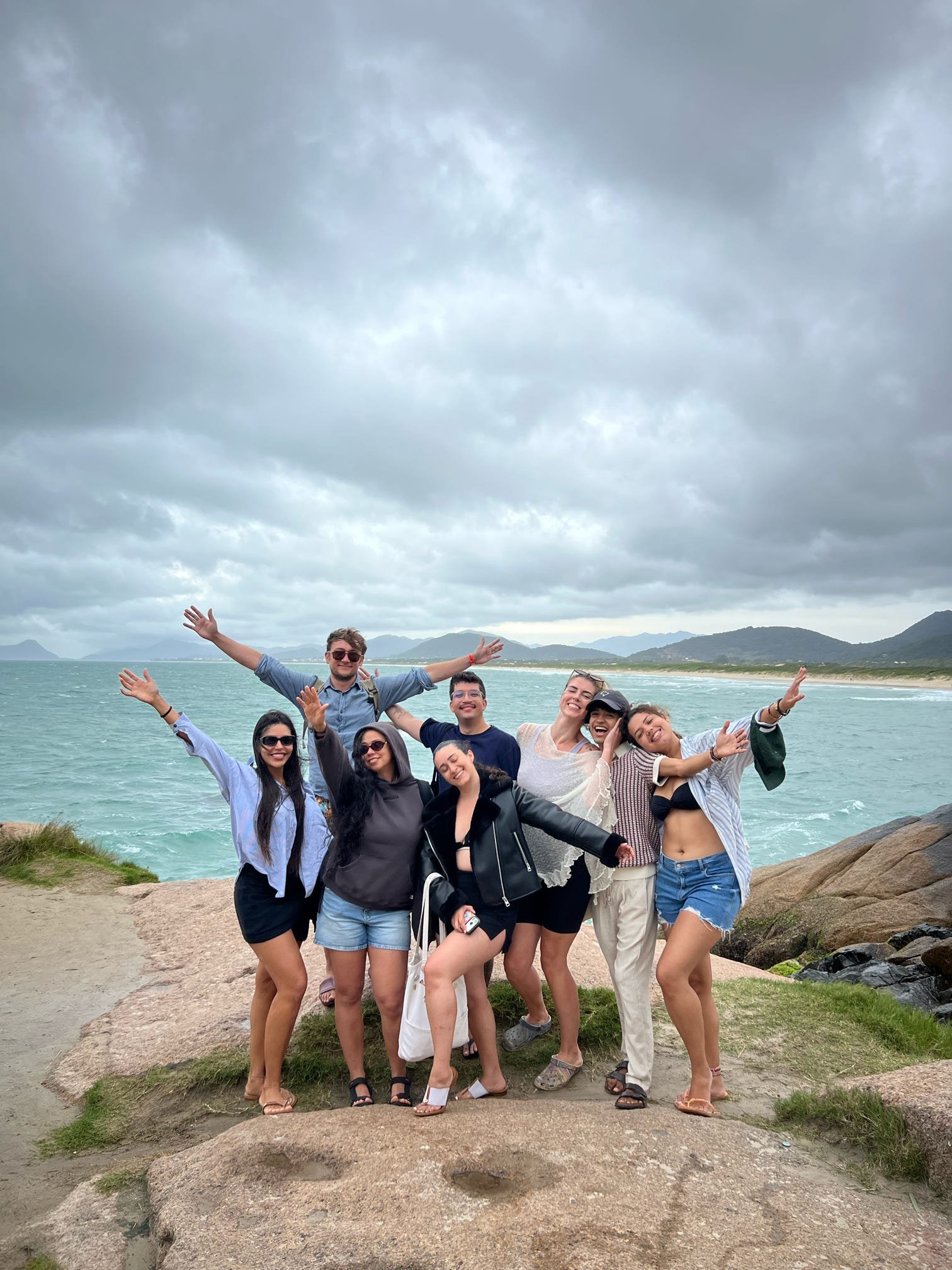 A group of smiling people posing on rocks near the water with mountains and cloudy sky in the background. Florianopolis, Brazil