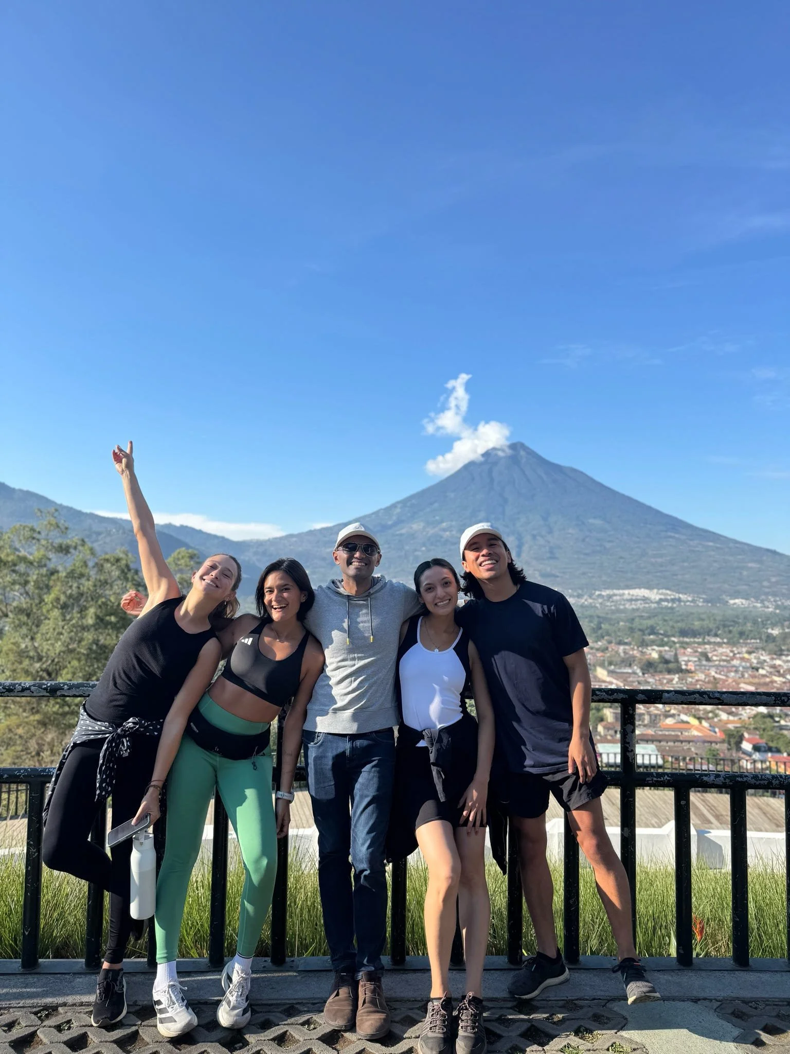 Group of five diverse friends smiling and posing with Agua Volcano, Antigua, Guatemala in the background, during daytime on clear weather.