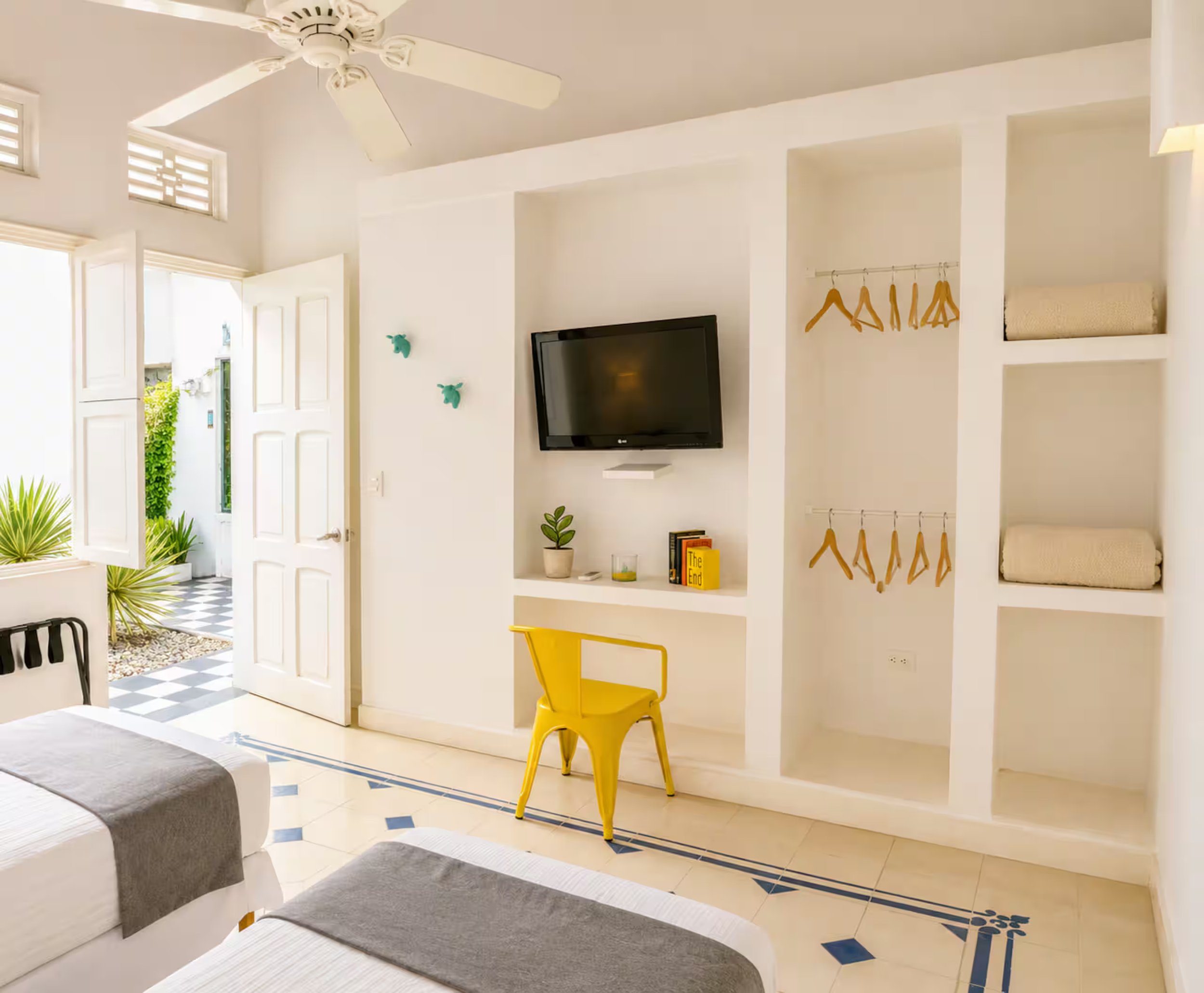 Interior of a bright, modern room with a white wall, a mounted black flat-screen TV, open shelving with books and a potted plant, yellow chair, and decorative wall hangings. An open door shows an outdoor area with checkered flooring and greenery.