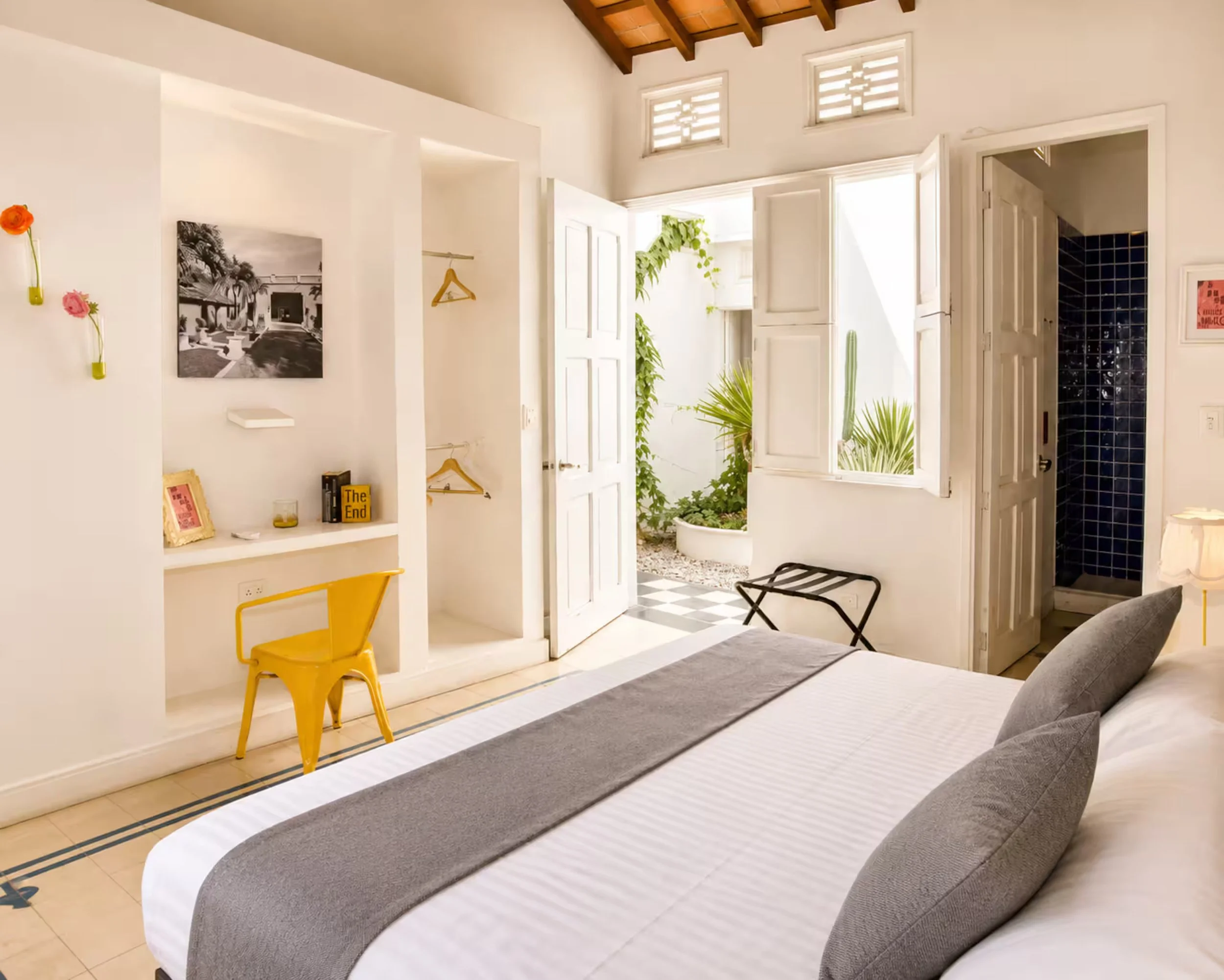 A bright bedroom with an open window featuring white shutters and a view of green plants outside. The room has a bed with a gray blanket, a gray chair, a yellow chair near a white wall, and wall art including a black-and-white photograph and small de