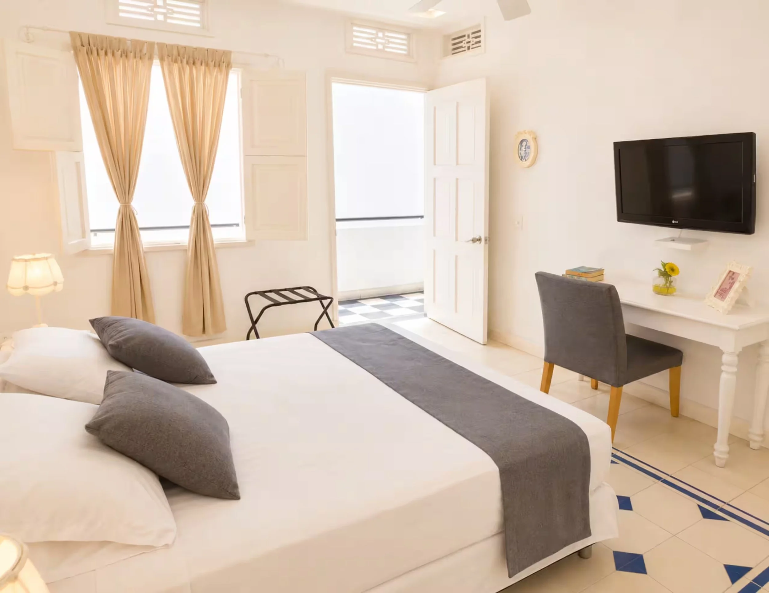 A bedroom with a bed covered with white sheets and gray pillows, beige curtains over windows, a wall-mounted flat-screen TV, a desk with books, a chair, and a small vase with a sunflower.