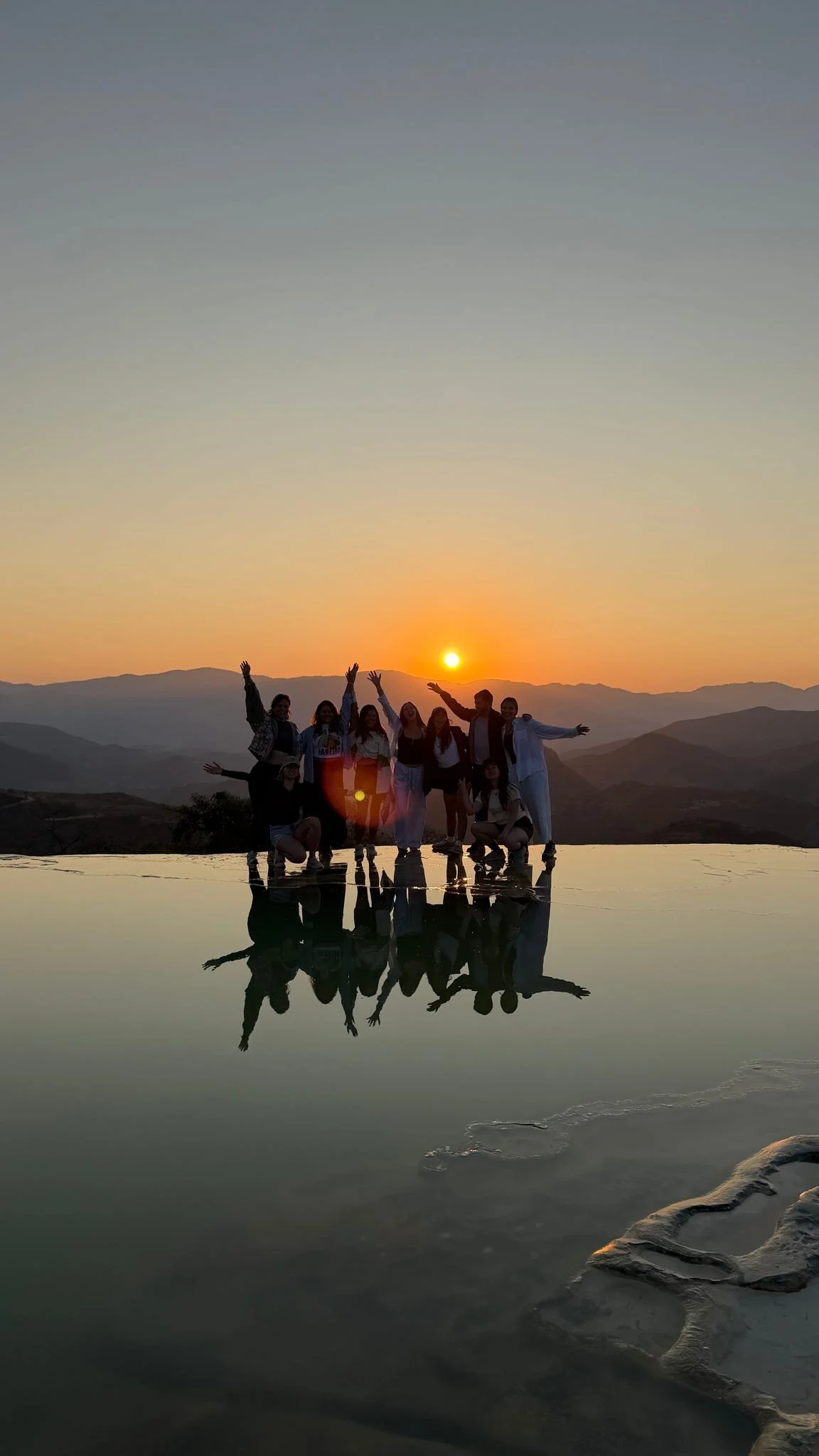 A group of people standing on a reflective surface at sunset with mountains in the background. They are raising their arms and smiling. Hierve el Agua, Oaxaca City, Mexico.