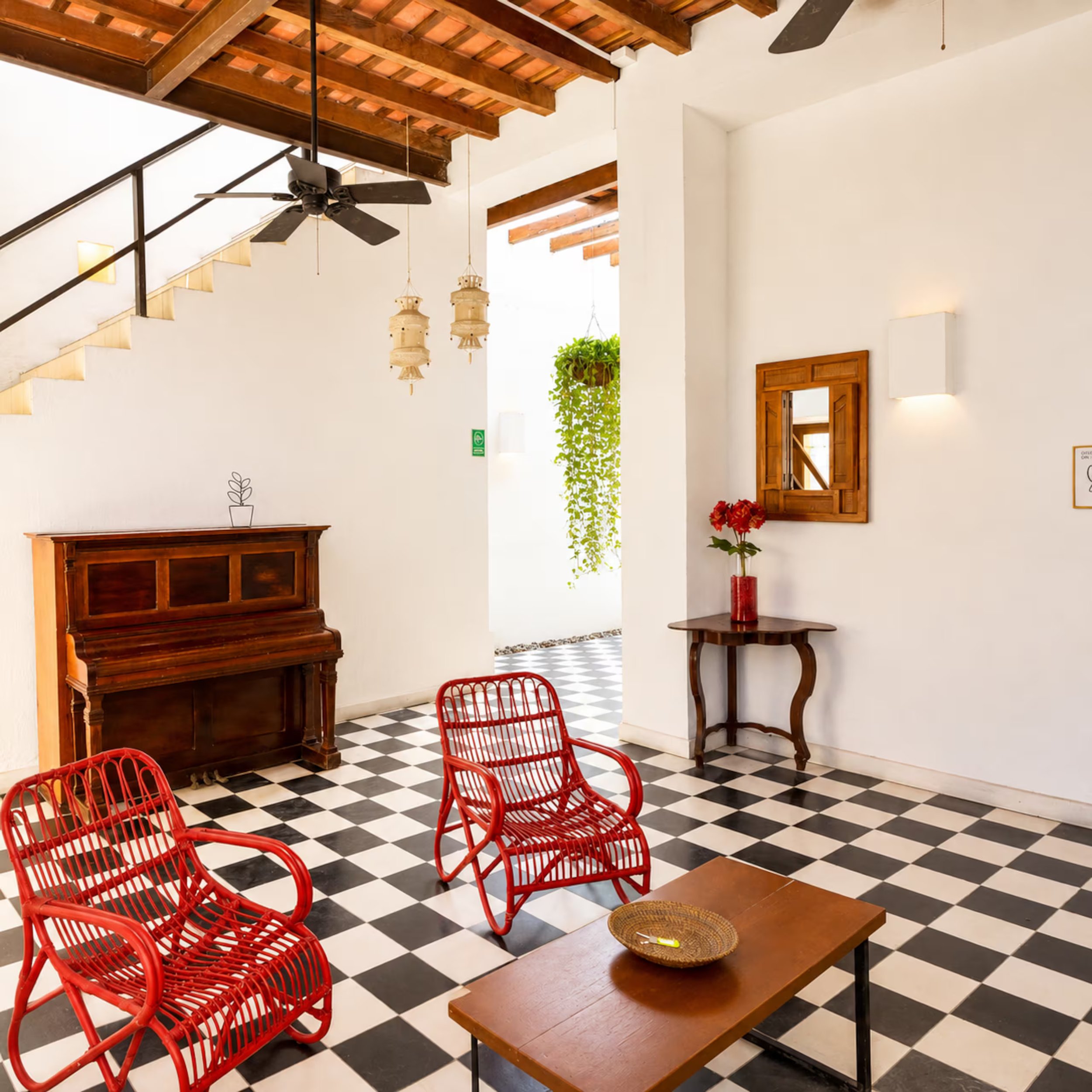 A cozy interior space with a white wall, wooden accents, a black and white checkered floor, red wire chairs, a wooden coffee table with a basket, a vintage wooden cabinet, hanging lanterns, and a ceiling fan with dark blades.