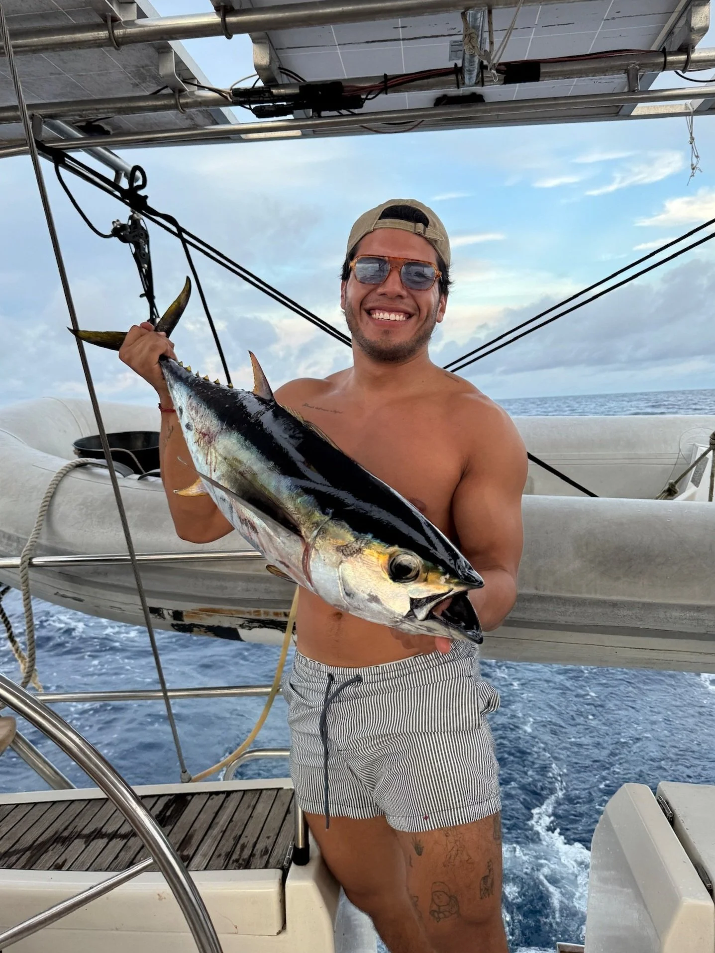 A man on a boat holding a large fish he caught, smiling at the camera with the ocean and sky in the background.