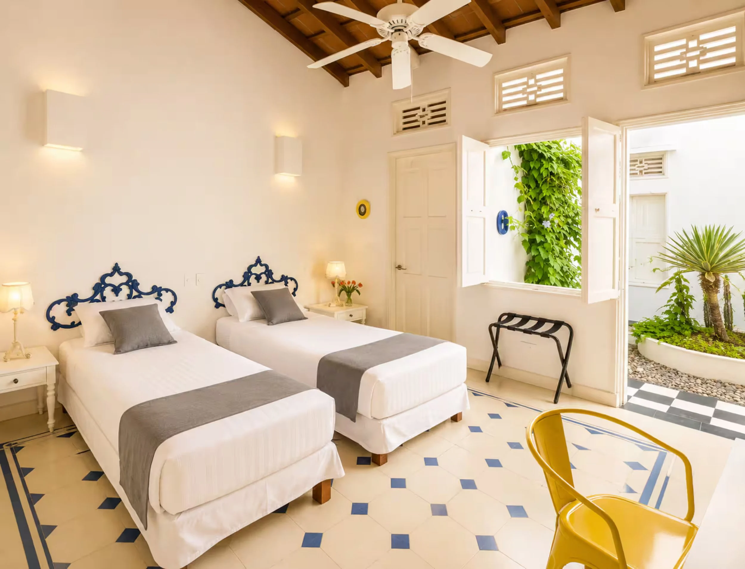 A bright bedroom with two white beds with blue decorative headboards, grey pillows, white nightstands with lamps and flowers, open windows with greenery outside, a ceiling fan, a black luggage rack, a yellow chair, and a checkered black and white til