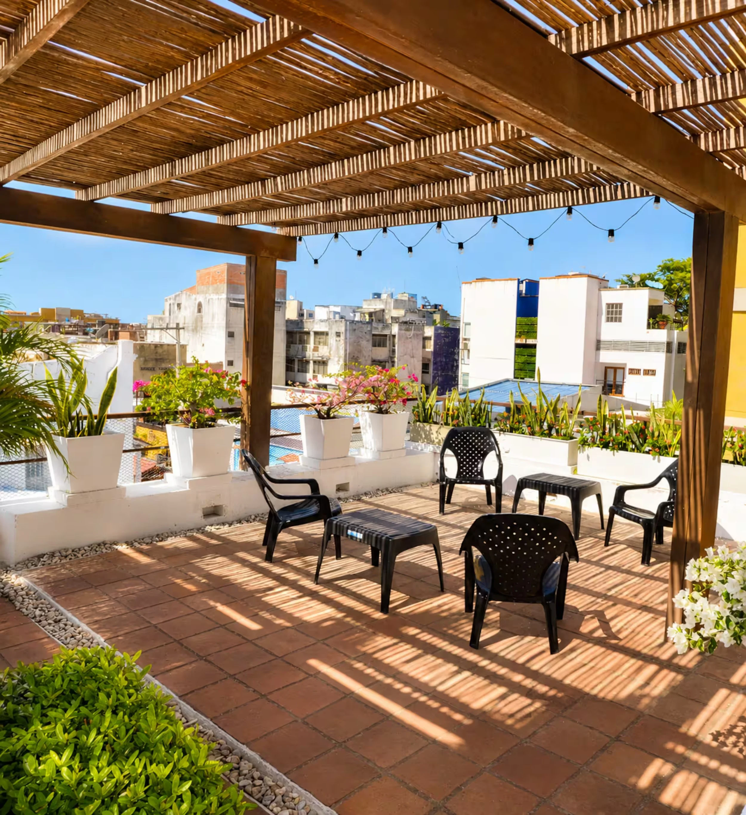 A rooftop terrace with a wooden pergola casting shadows over a seating area, potted plants, and city buildings in the background under a clear blue sky.