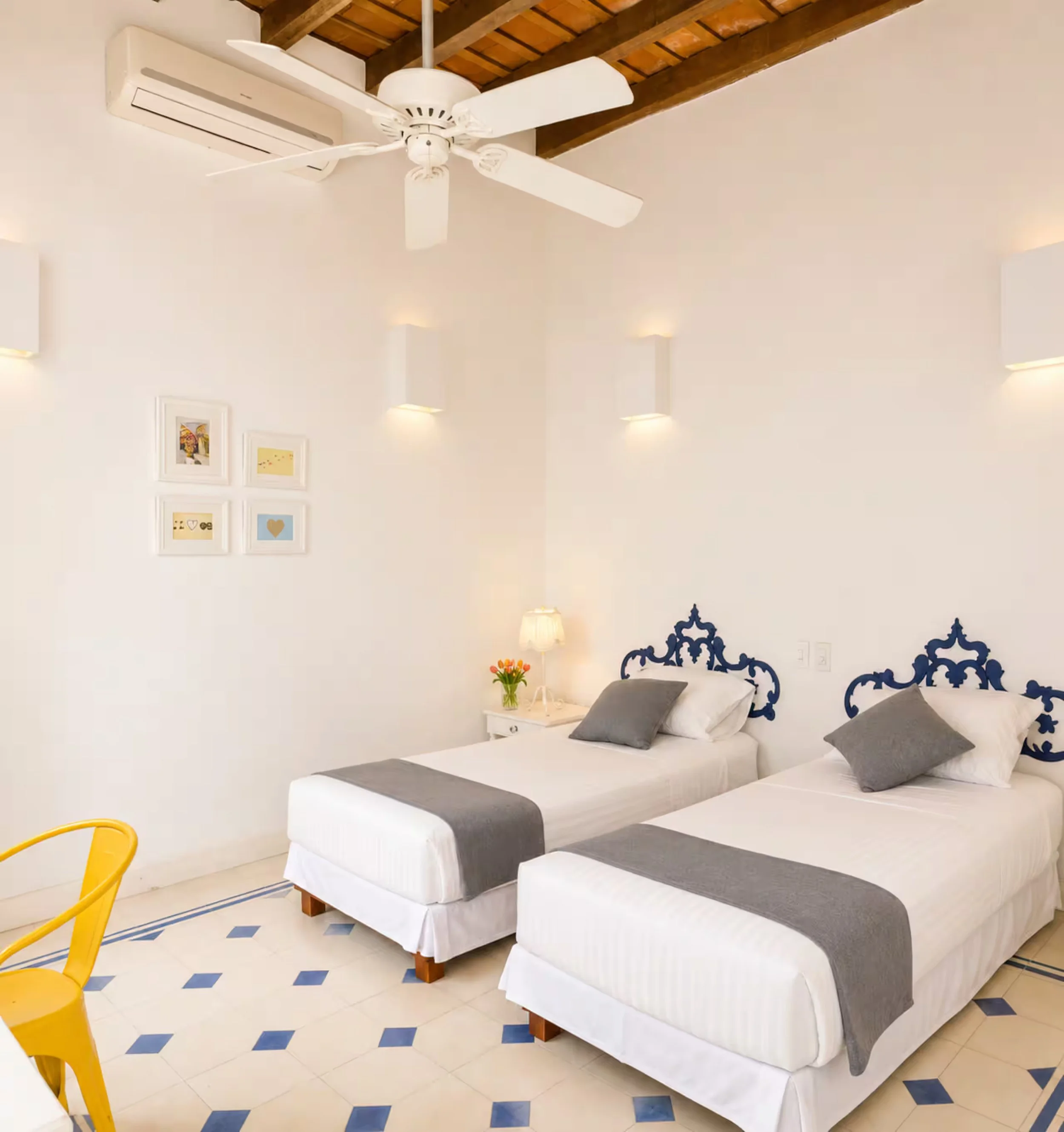 A bedroom with a white bed that has navy and white striped bed linens and gray pillows. There is a white nightstand with a vase of orange and yellow flowers, a white lamp, and framed artwork on the wall. The room has white walls with soft lighting, a