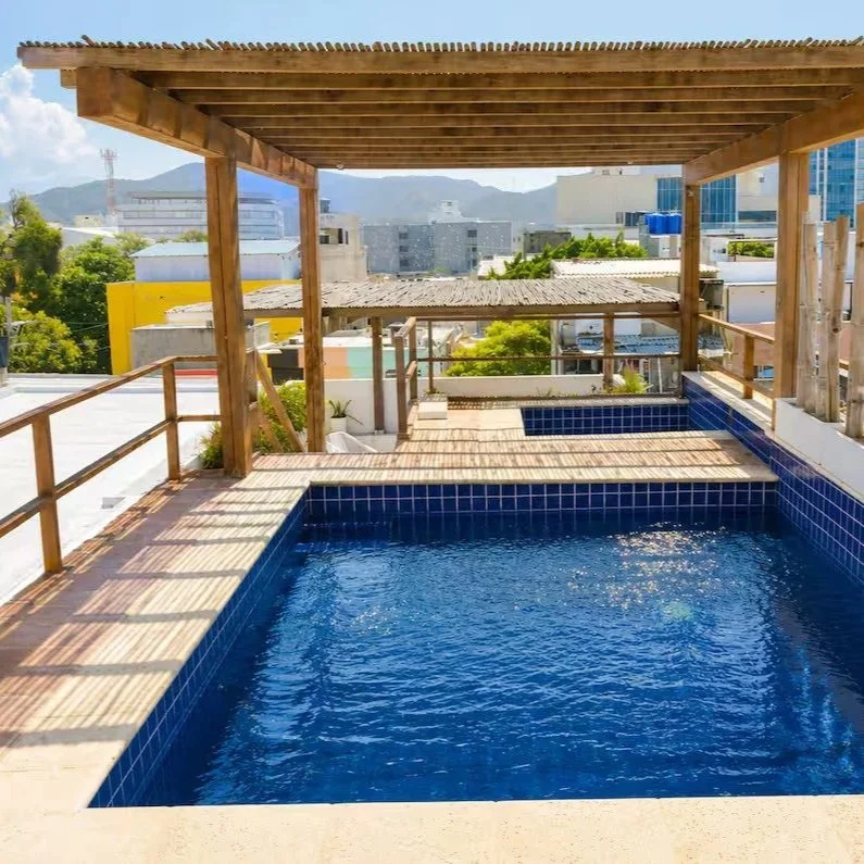 Rooftop swimming pool with wooden deck and pergola, overlooking city buildings and mountains in the distance