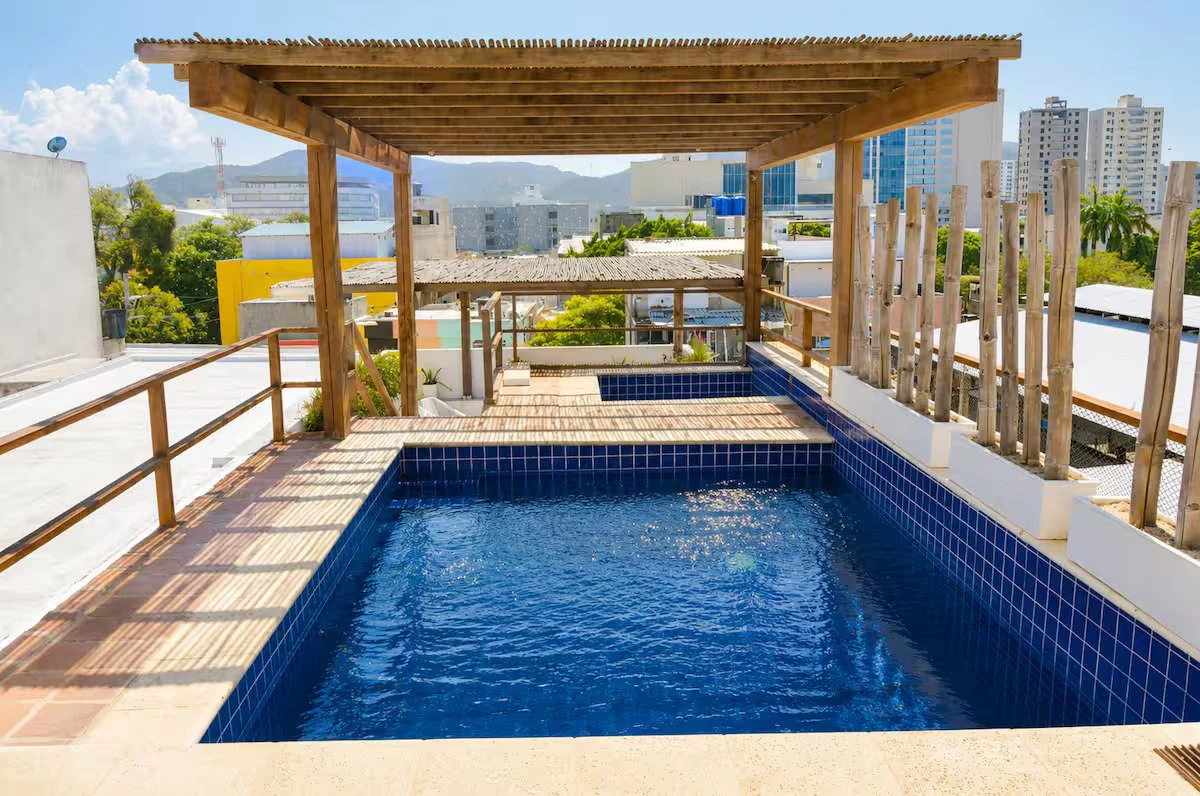 Rooftop swimming pool with blue tiles, wooden deck, and a wooden pergola, overlooking city buildings and distant mountains under a partly cloudy sky.