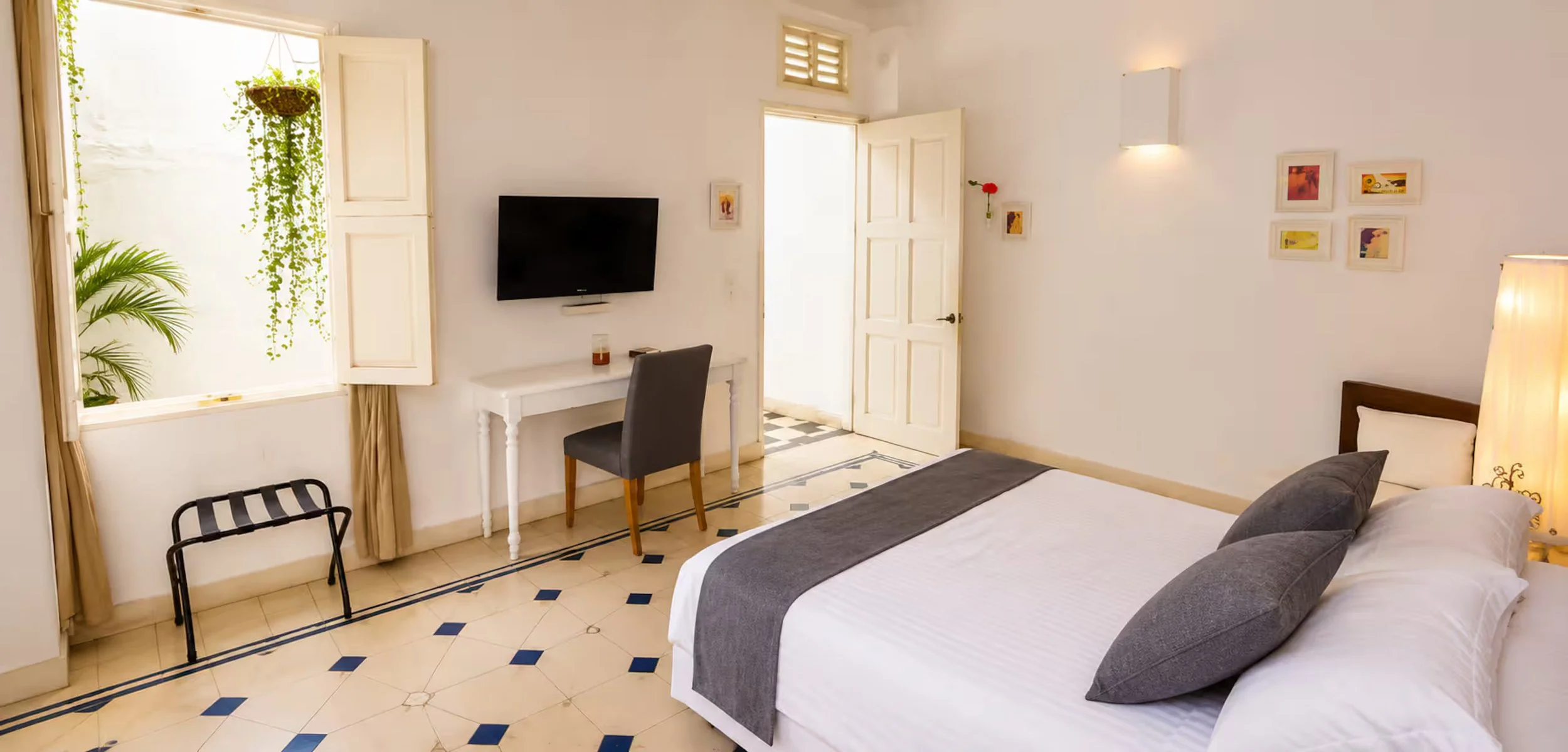Sunlit hotel room with a double bed, pillows, and a gray blanket, a wall-mounted TV, a small white desk with a chair, a window with open shutters, indoor plants, and wall decor.