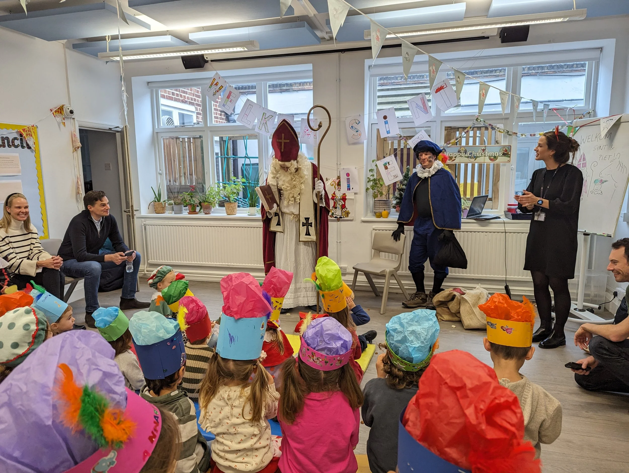 Kinderen kijken naar een Sinterklaas- en Zwarte Piet-vertoning in een klaslokaal, met volwassenen die erbij staan, versierd met feestelijke slingers en papieren mutsen.