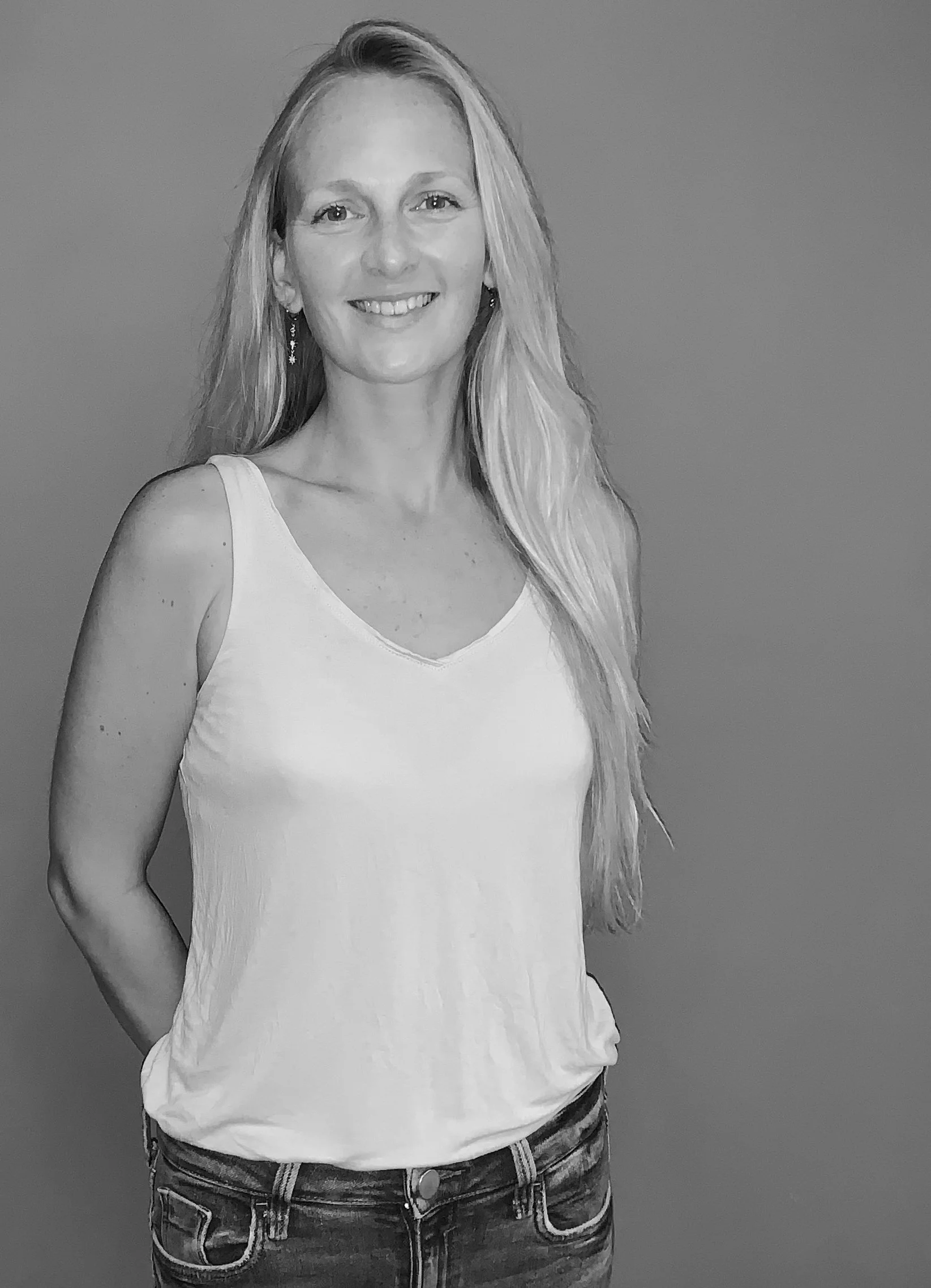 A smiling woman with long blonde hair in a sleeveless white top and jeans, standing against a plain background.