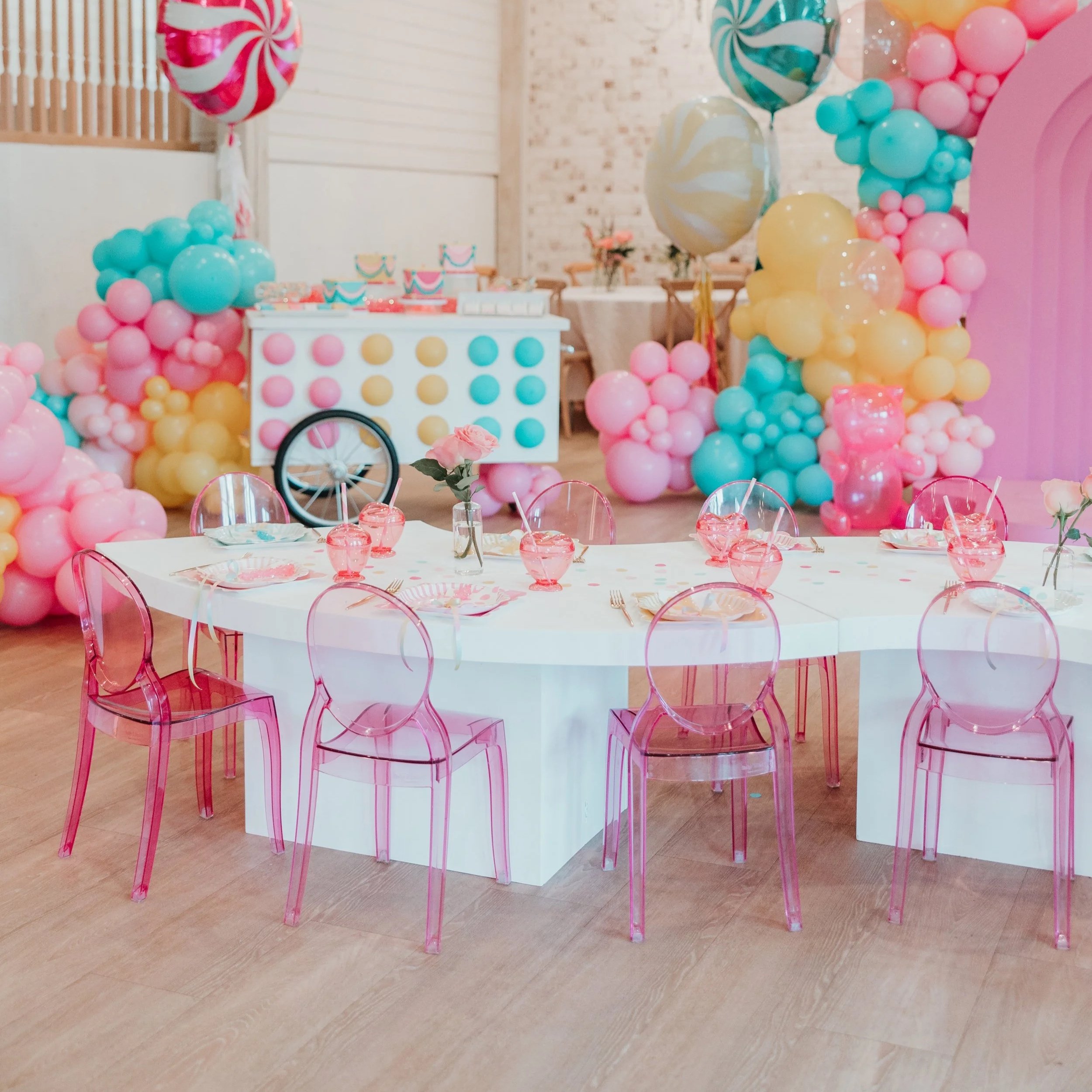 Decorated birthday party table with pink and transparent chairs, pink glasses, and a balloon arch in pastel colors including pink, blue, and yellow, with a backdrop of balloons and a decorated dessert table.