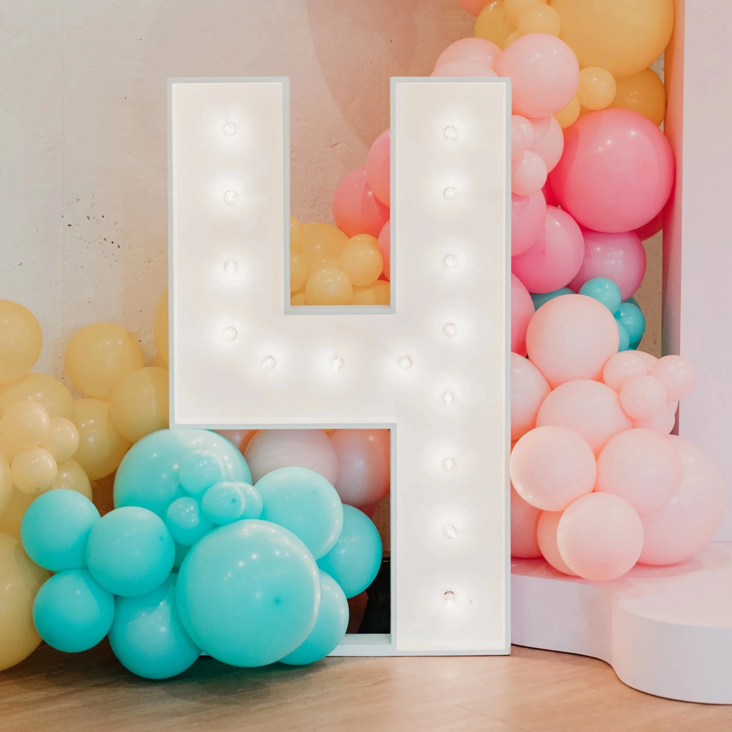 A large white letter 'H' with built-in lights, surrounded by pink, yellow, white, and blue balloons in various sizes.