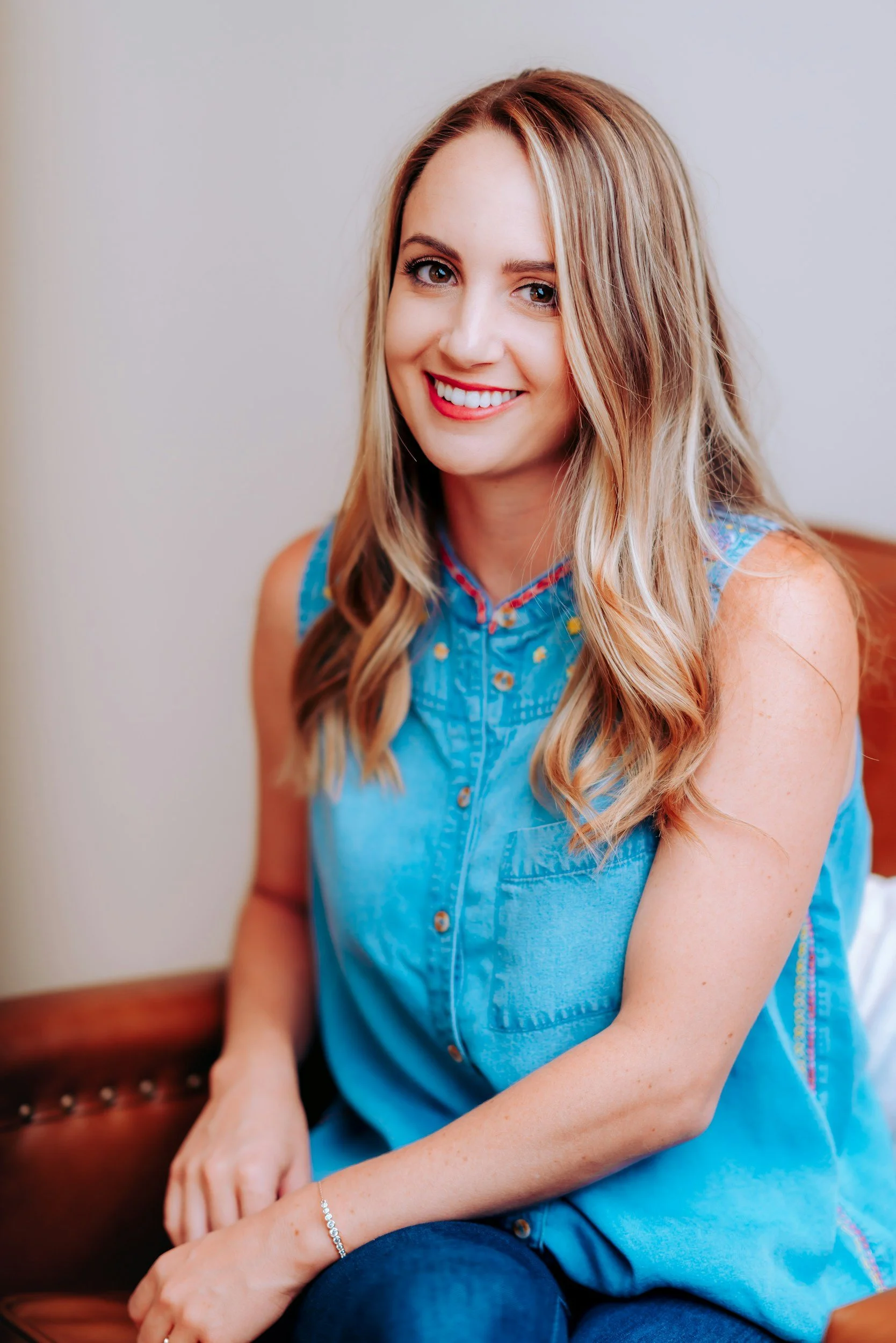 A woman with long blonde hair and blue eyes, smiling and wearing a blue sleeveless denim shirt with brown buttons, sitting on a leather chair.