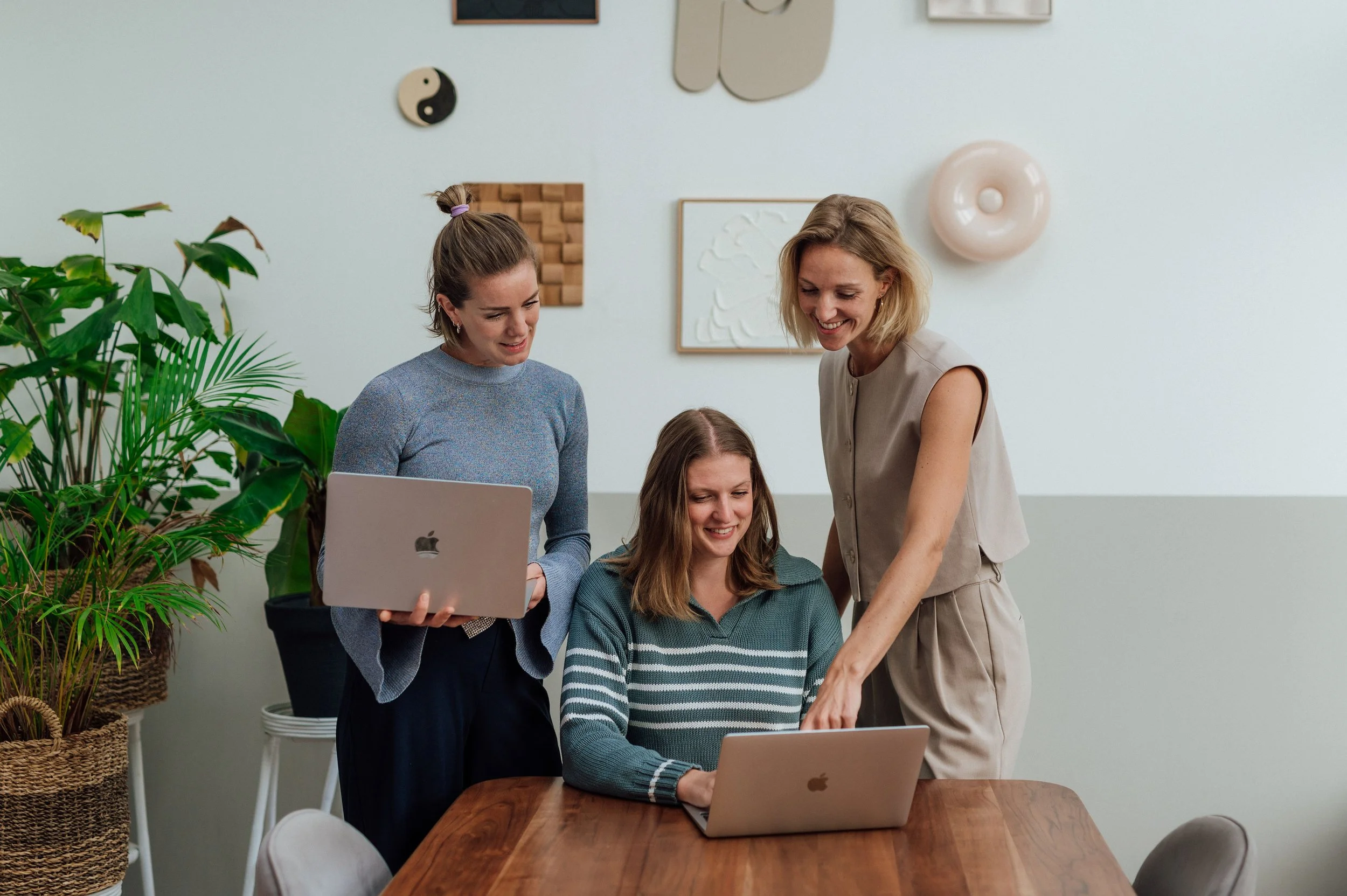 Drie vrouwen werken samen aan laptops in een vergaderruimte met groene planten en muurdecoraties.