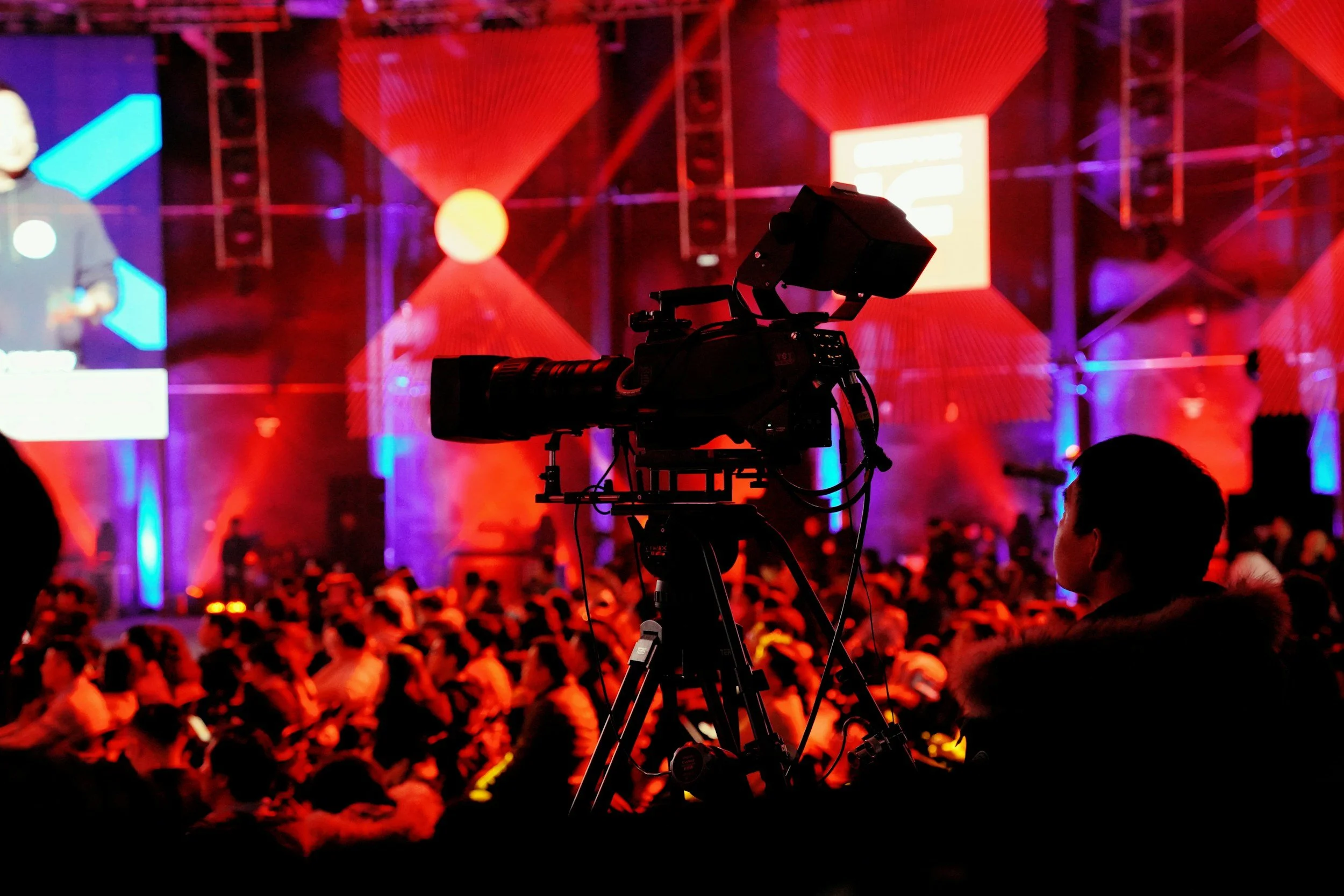 Conference room with a man giving a presentation at a podium, projected slides, and camera equipment recording the event.