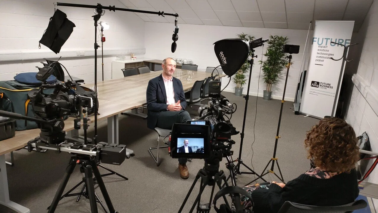 Laptop on desk displaying a virtual meeting with multiple participants, in a classroom setting, with a smartphone beside it and a woman wearing a mask seated at another desk.