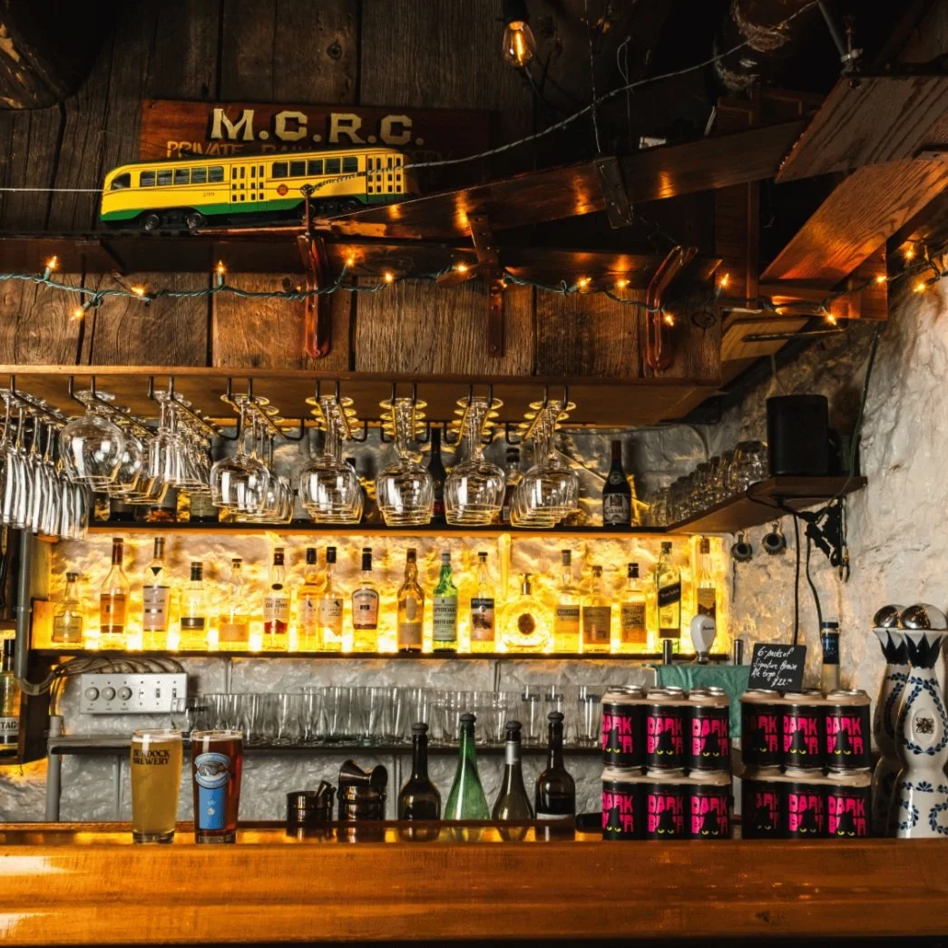 Interior of a pub with a wood shelf displaying liquor bottles, hanging glassware, and a decorative model train on a wooden beam.