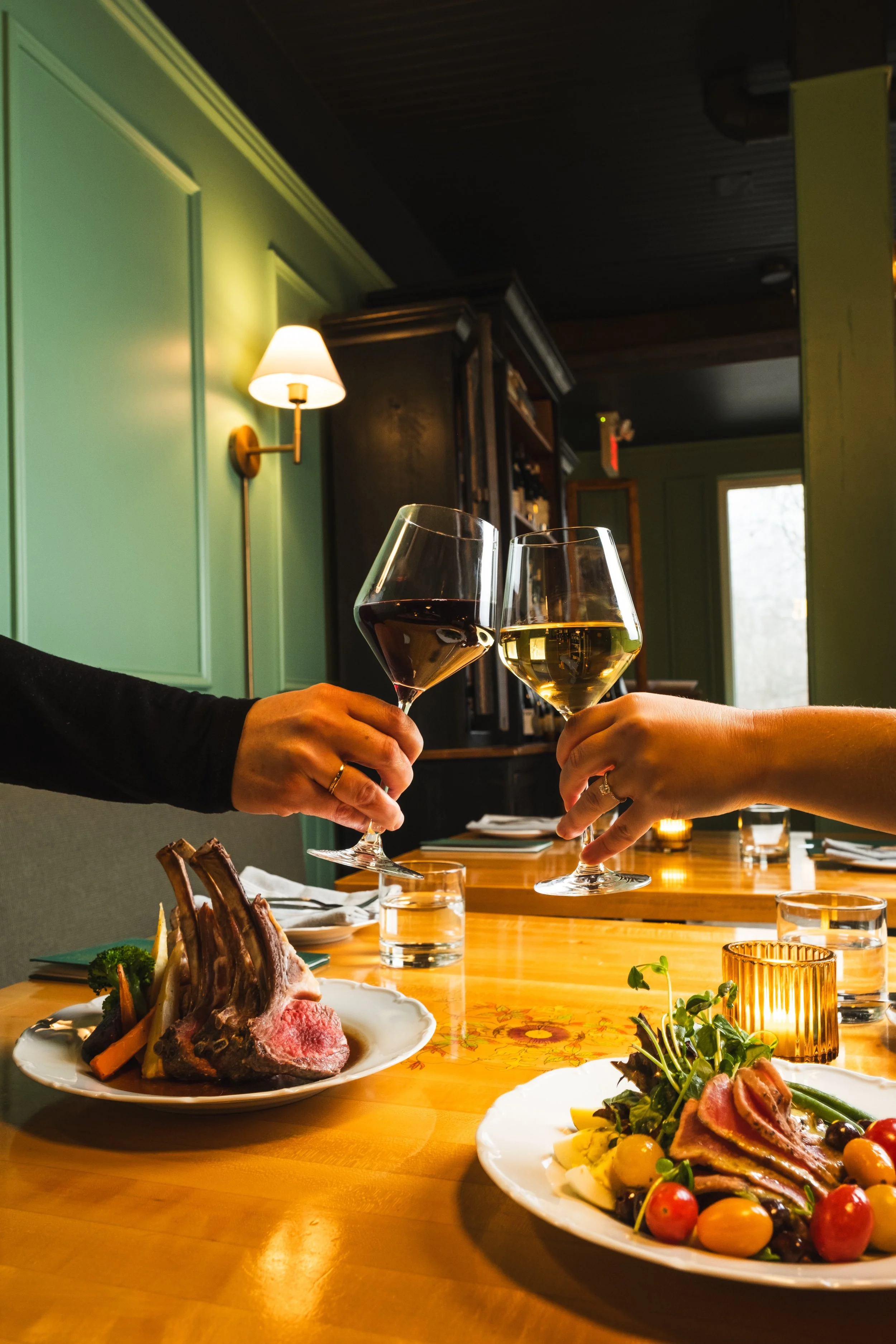 Two people clinking glasses of red and white wine at a dinner table with plates of food, including lamb chops and a salad, in a warmly lit restaurant.