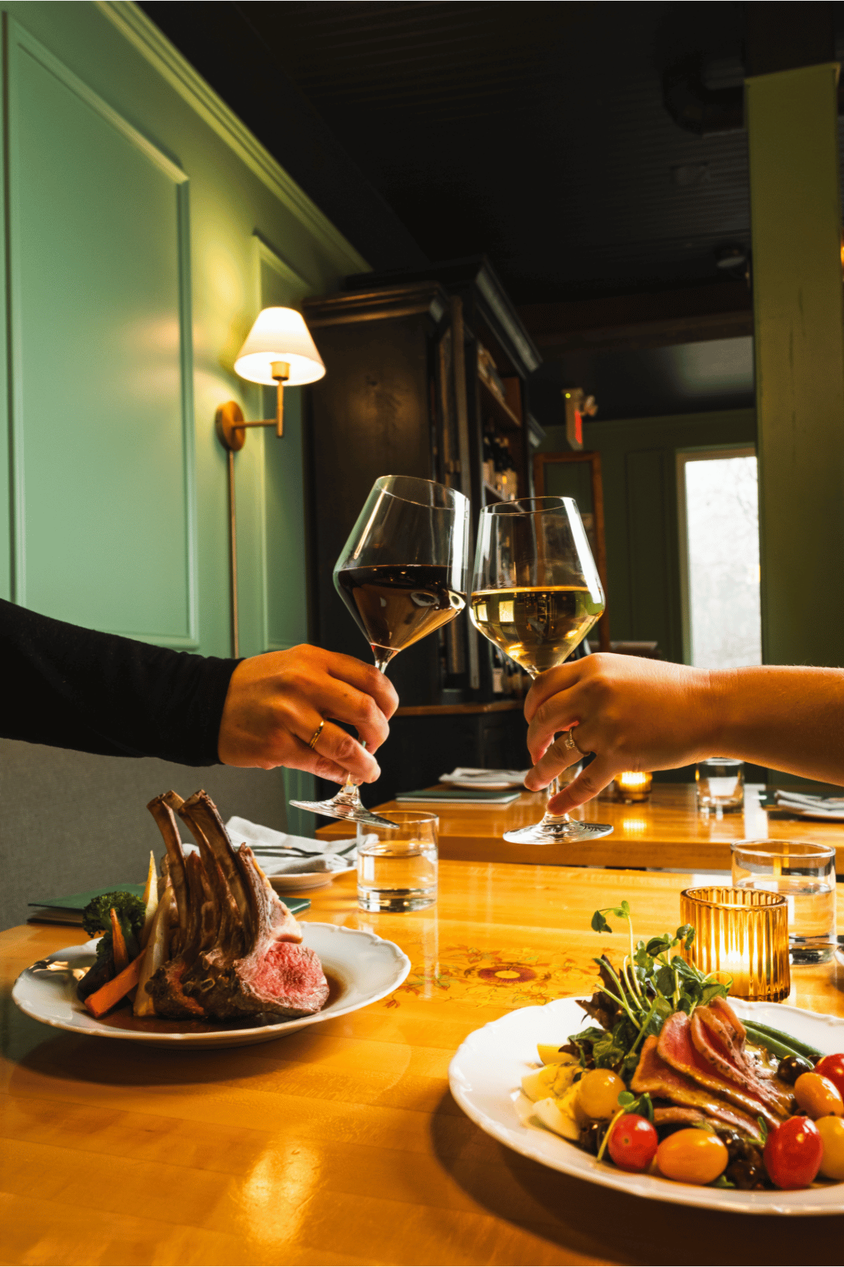Two people clinking glasses of white wine at a dinner table with plates of lamb chops and salad, in a cozy restaurant setting.