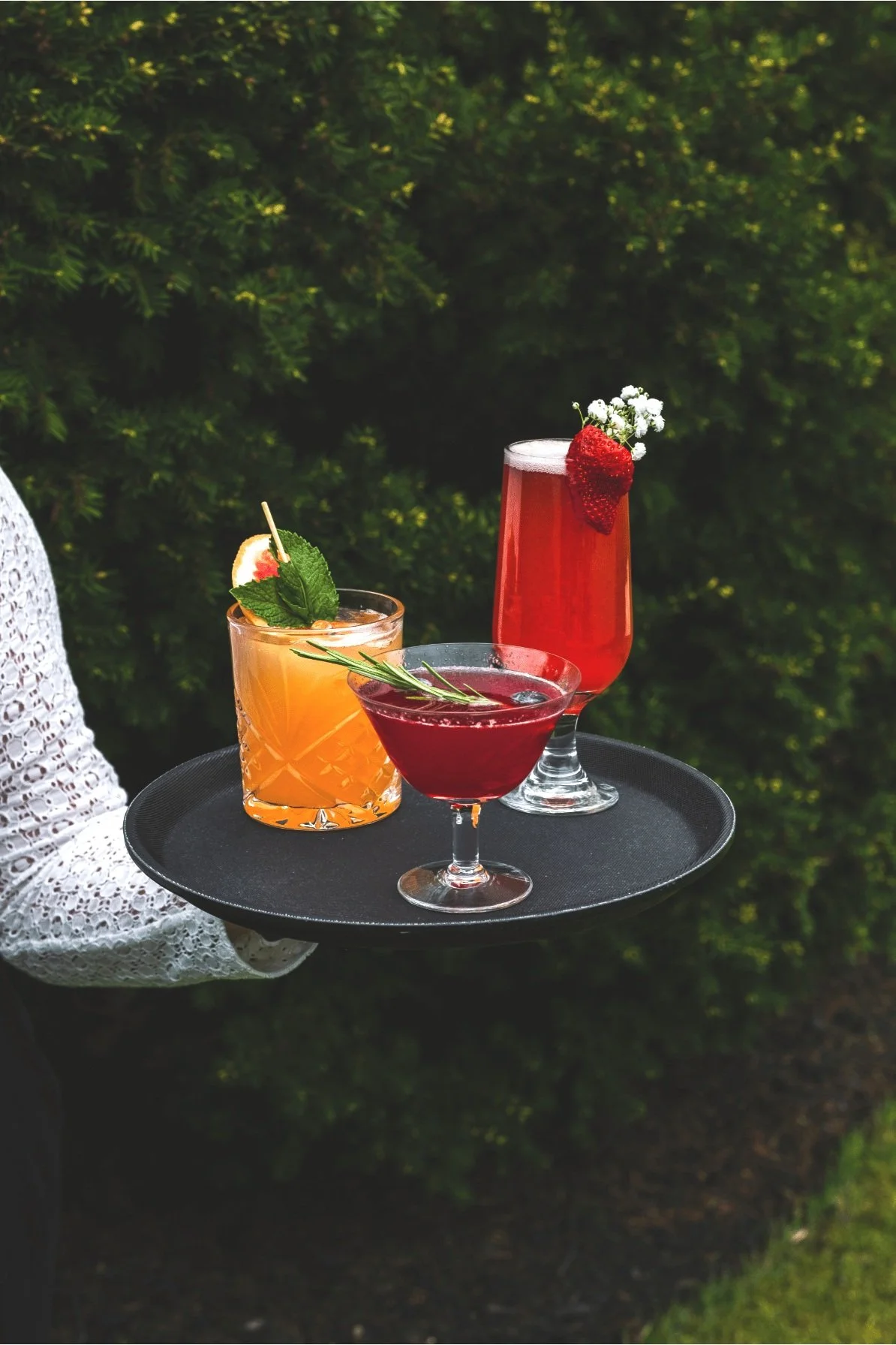A person holding a black tray with three colorful mocktails outdoors against green foliage. The cocktails are garnished with fruit, herbs, and flowers.