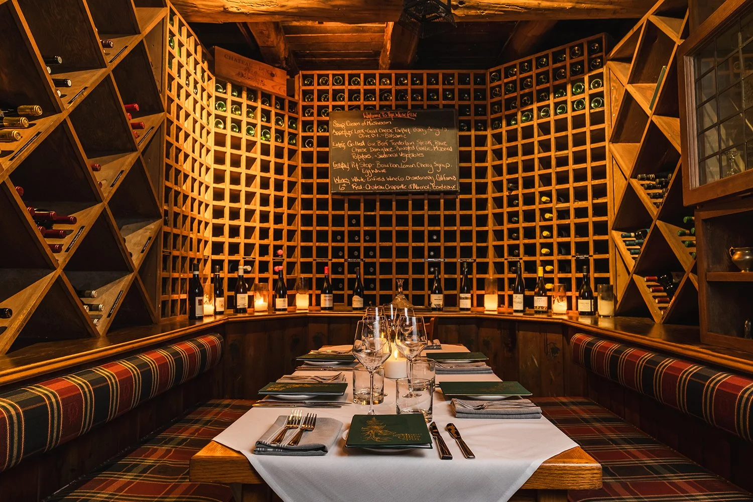 A cozy restaurant wine cellar with wooden wine racks filled with bottles surrounding a dining table set with plates, silverware, glassware, candles, and lanterns.