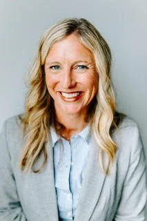 A white woman with blue eyes and long, dark blonde hair, wearing a light grey blazer and a light blue, button down shirt.