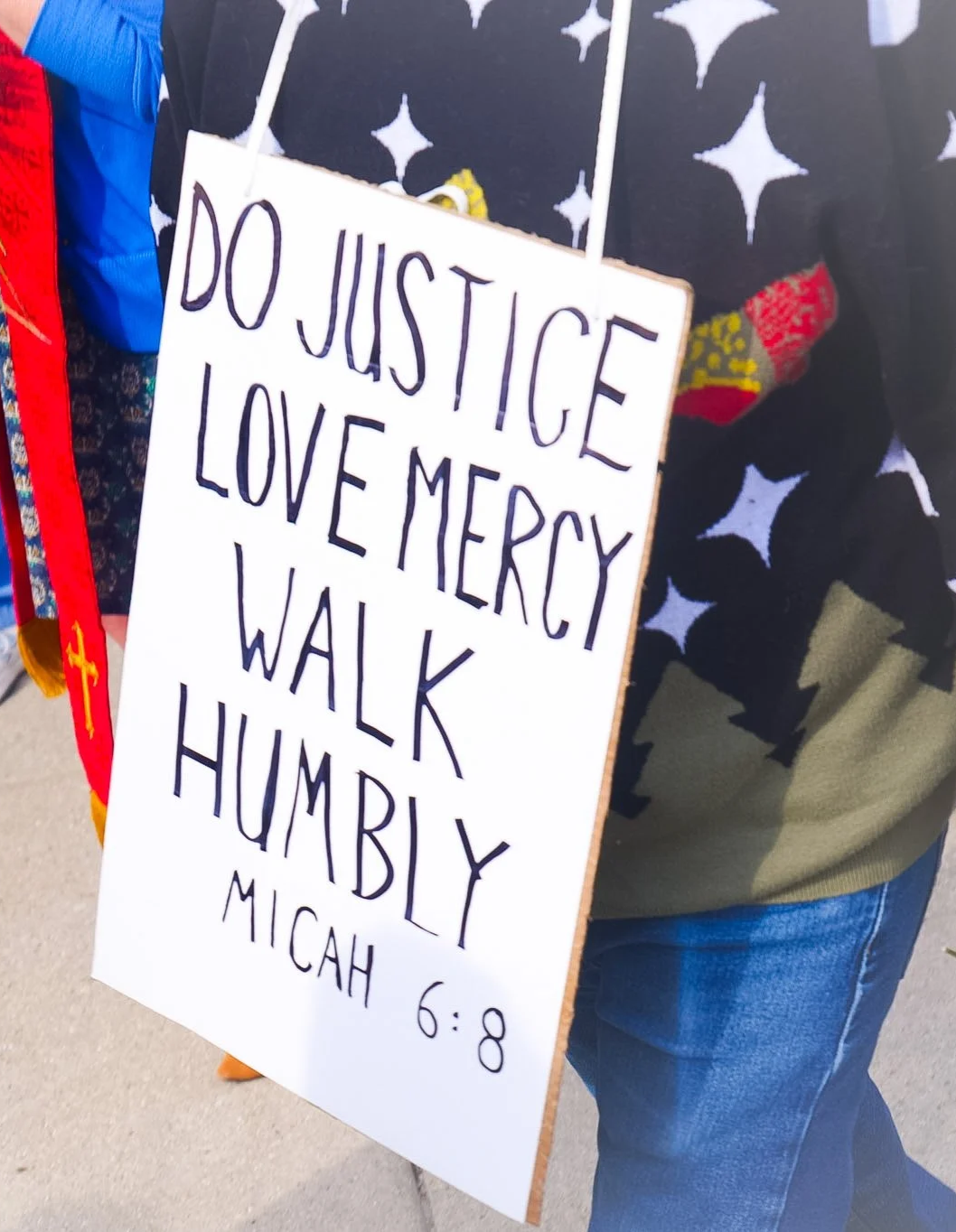 A large, white sign with black text reading, "Do Justice. Love Mercy. Walk Humbly. Micah 6:8" is worn by someone wearing jeans a a sweater, walking through the streets with a crowd.
