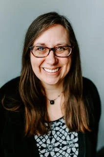 A white woman with long brown hair, wearing tortoiseshell glasses, a necklace, a black cardigan, and a black and white, patterned top.