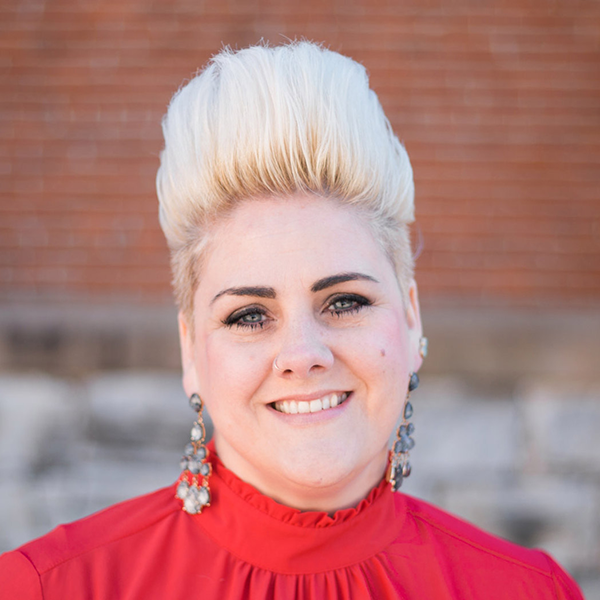 A photo of a smiling woman with pale peach skin and very tall, icy blonde hair. She is wearing a red shirt and shiny, dangling earrings.