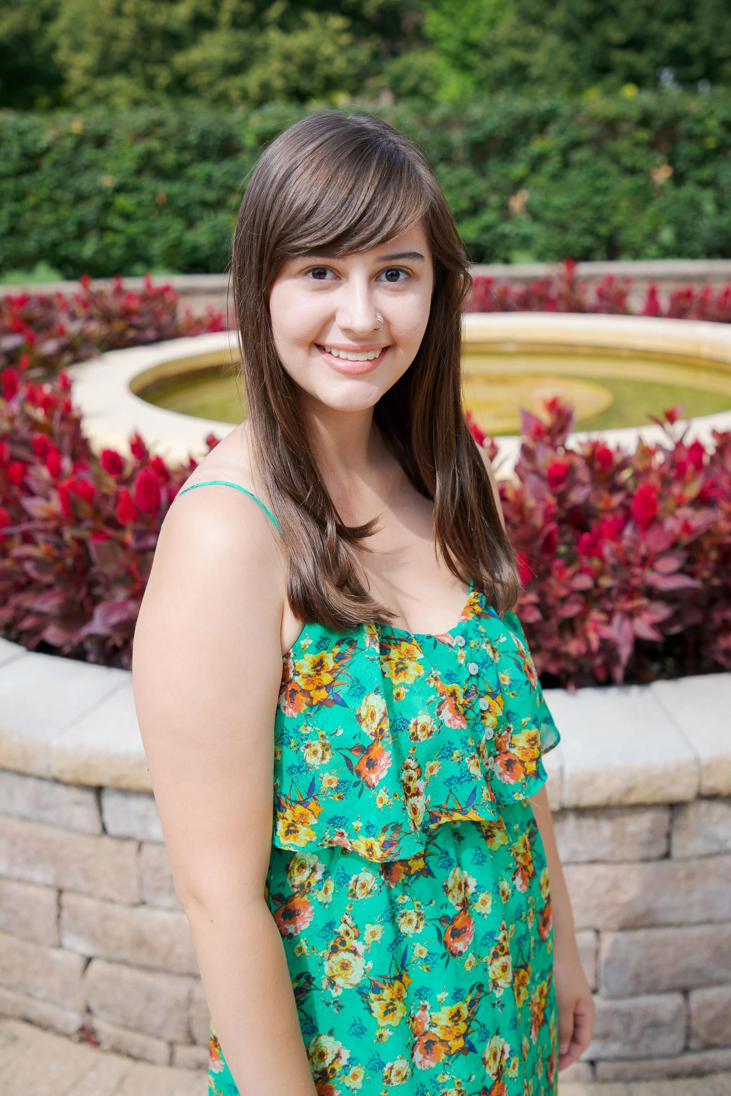 Photo of Kole Torres, a young, light-skinned woman with long brown hair and brown eyes, wearing a colorful dress, standing in front of a planter with dark red plants and green shrubs in the far background.