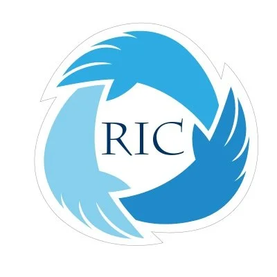 The logo of the Racine Interfaith Coalition – black text in the center reads, “RIC,” surrounded by figurative, wing-like hands in a triangle, in shades of light blue, seeming to hold on to each other.