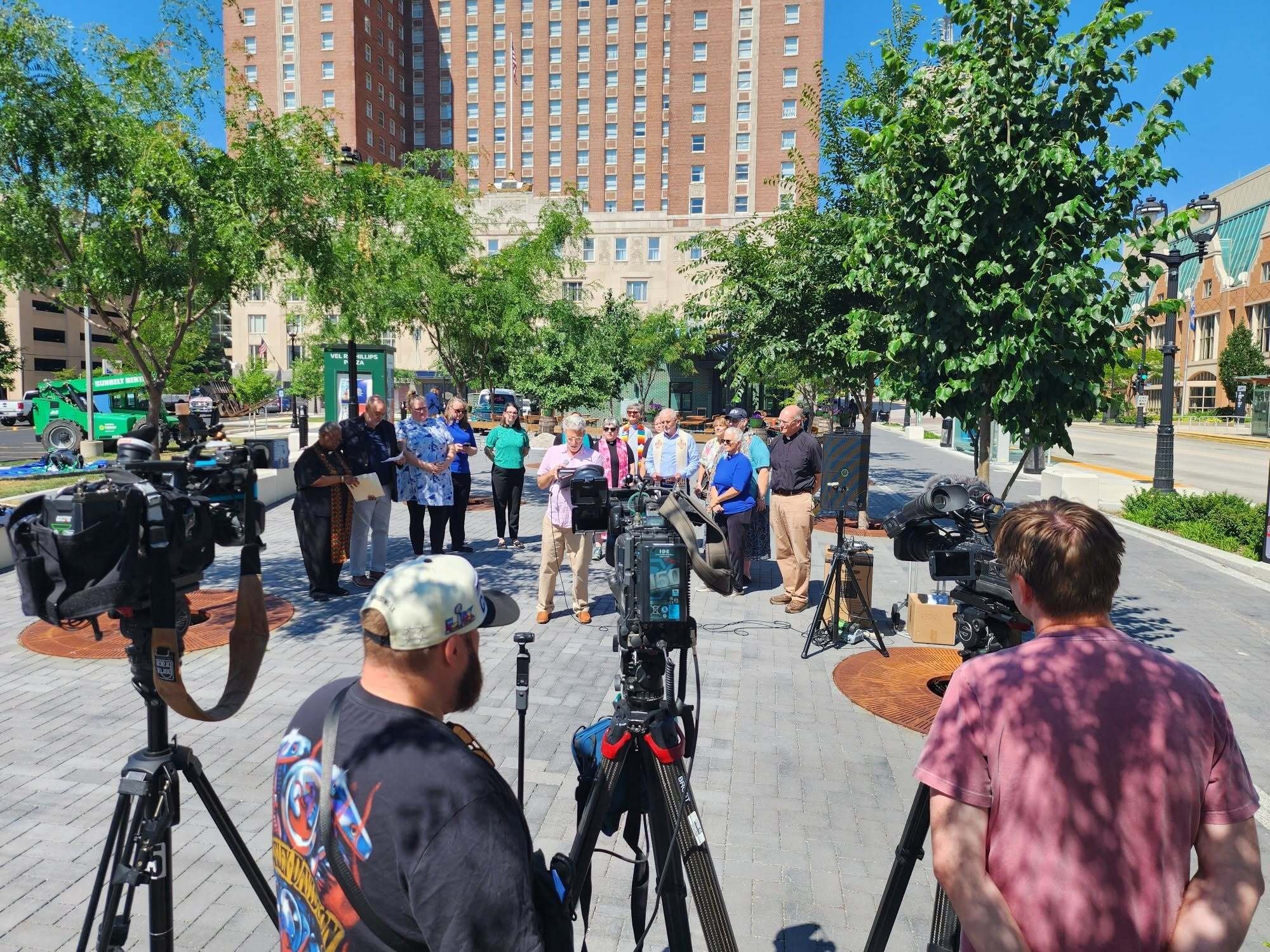 Clergy and faith leaders gather around a microphone at Vel Phillips Plaza in Milwaukee. In the foreground are several media cameras and camera operators. Photo credit: American Baptist Churches of Wisconsin.