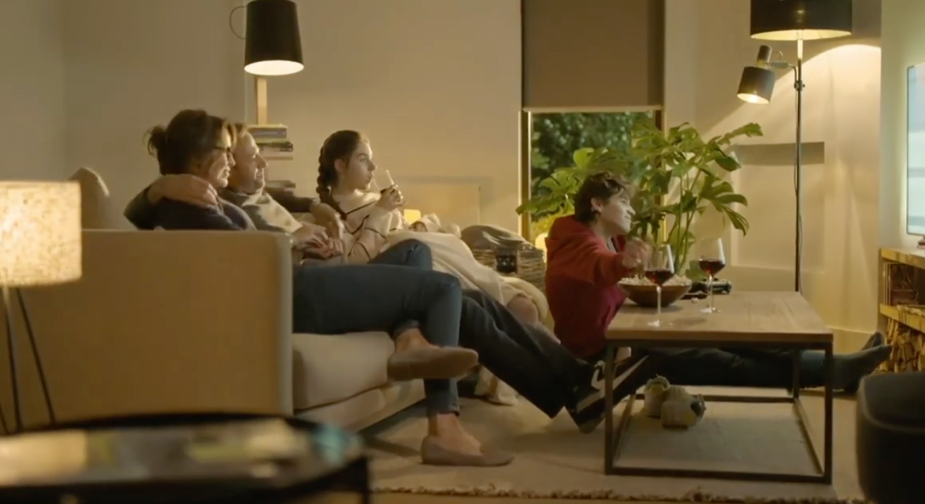 A group of four people sitting on a couch and floor watching television in a cozy living room, with a woman holding a glass of red wine and a man and woman embracing after having their Omni Smart Homes System Installed