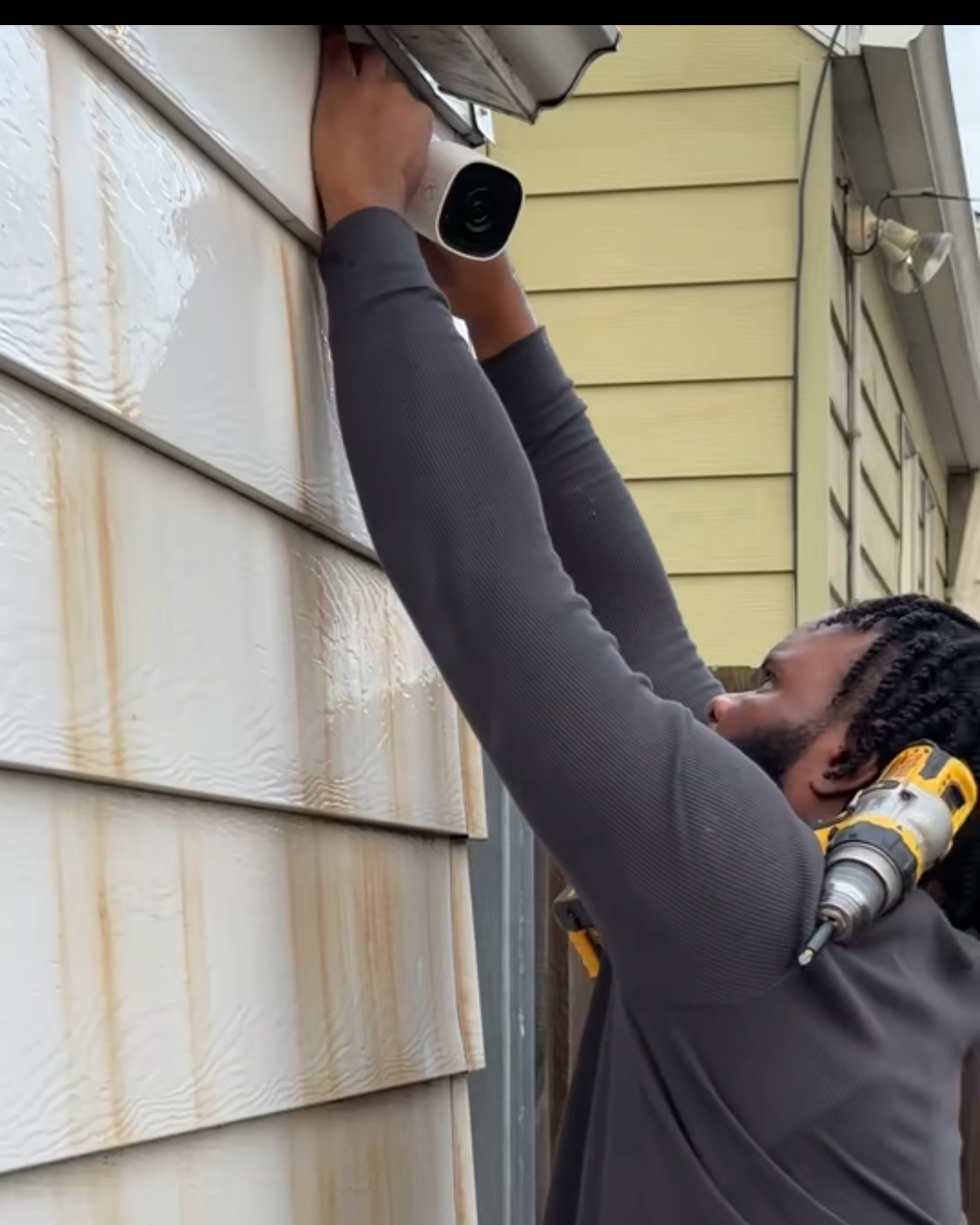 A man installing an alarm.com smart security camera with audio, video analytics notifications, and 24/7 recording on the exterior wall of a house.